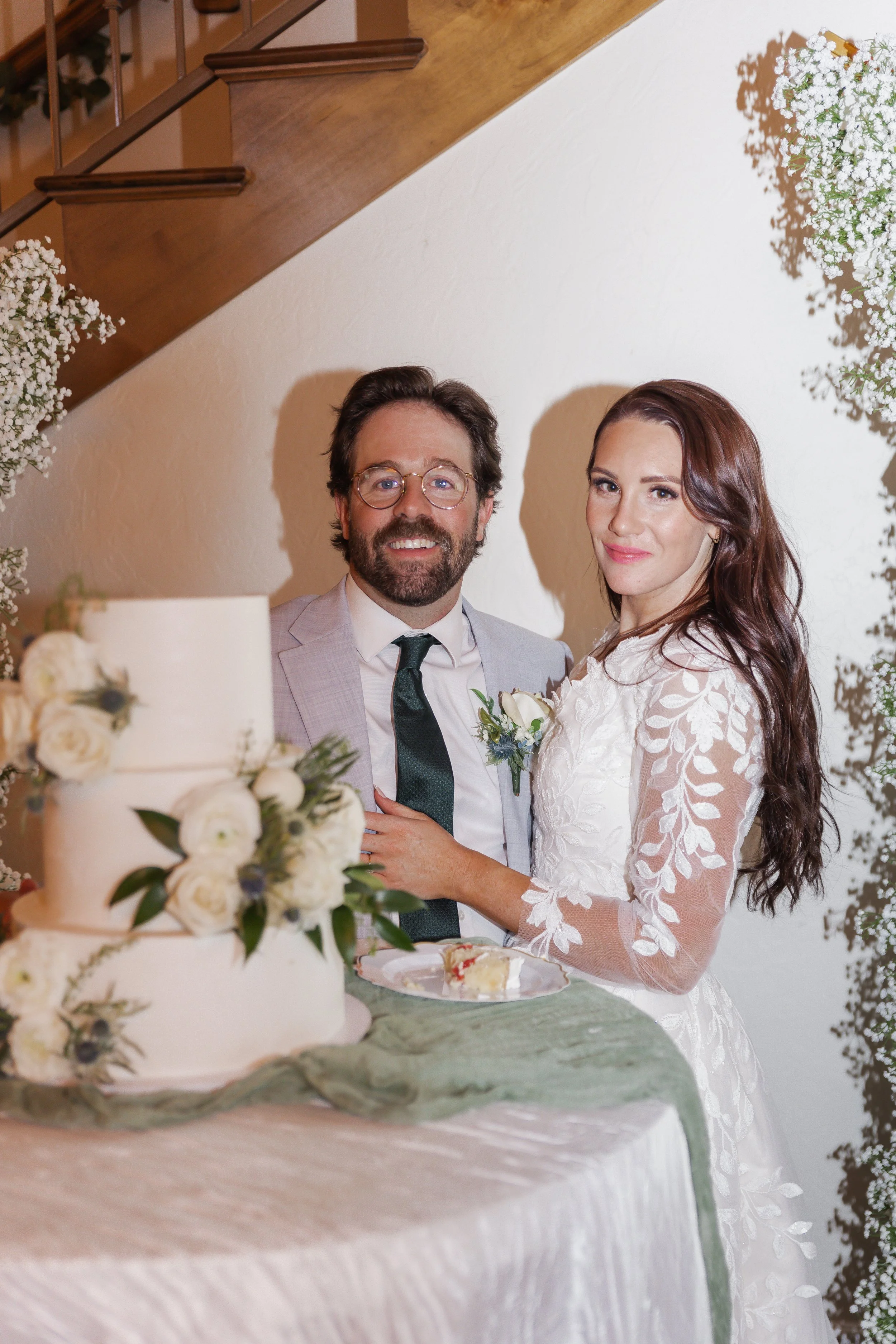 A bride and groom celebrating their wedding, standing behind a wedding cake decorated with white flowers. The bride has long wavy hair and is wearing a white lace wedding dress, while the groom has glasses, a beard, and is dressed in a light gray sui