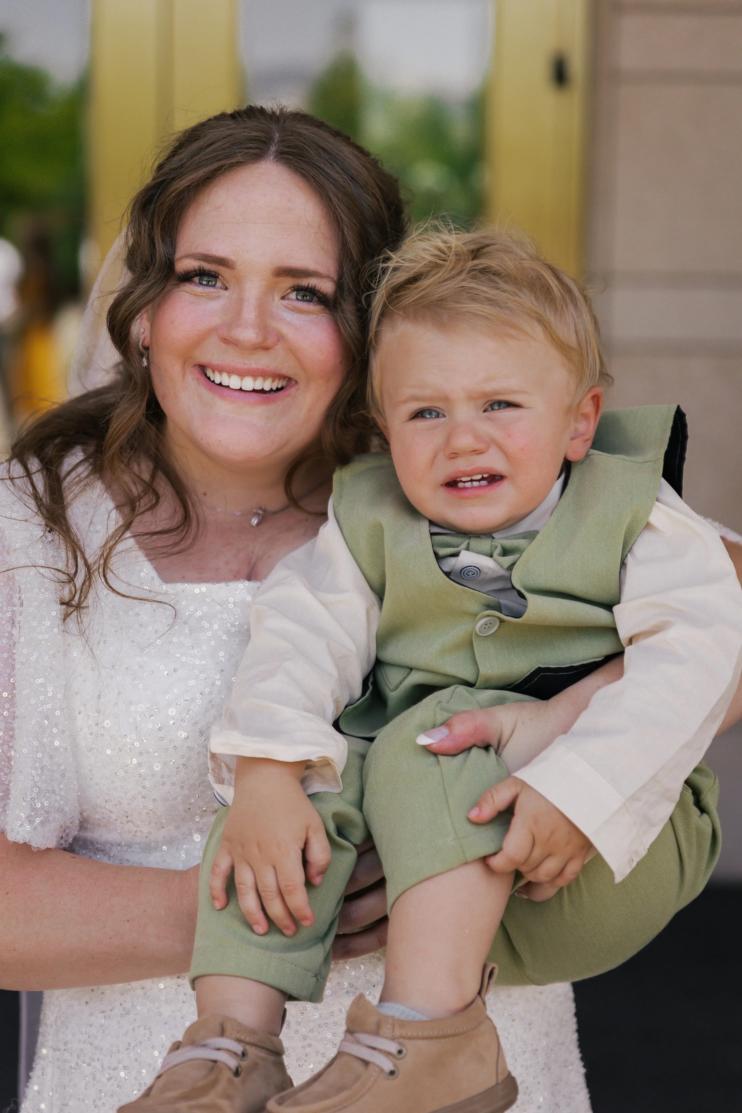 A woman in a white dress smiling, holding a young boy in green and beige formal clothes with a confused expression.