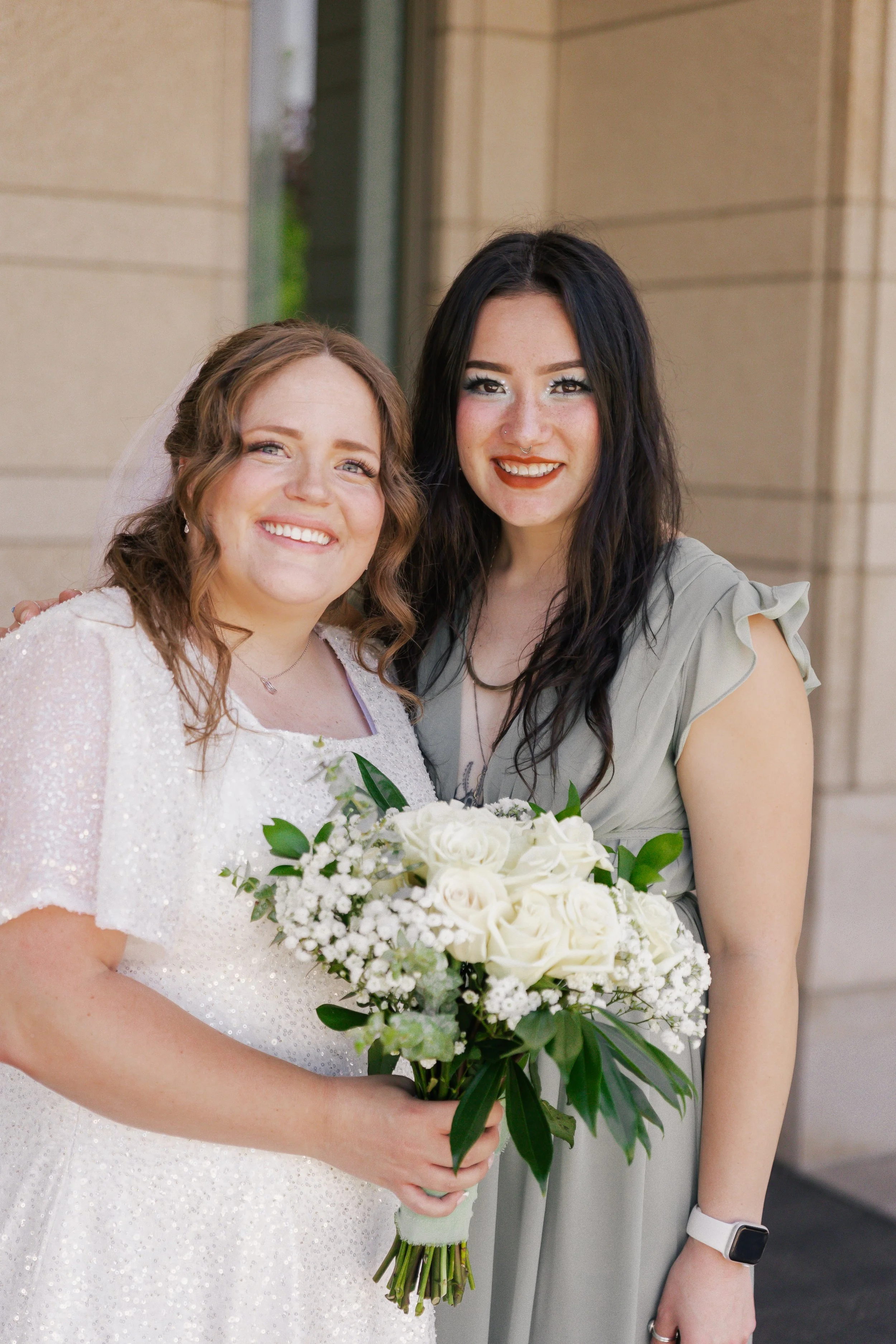Two women posing outside; one in a wedding dress holding a bouquet, and the other in a light green dress, smiling at the camera.