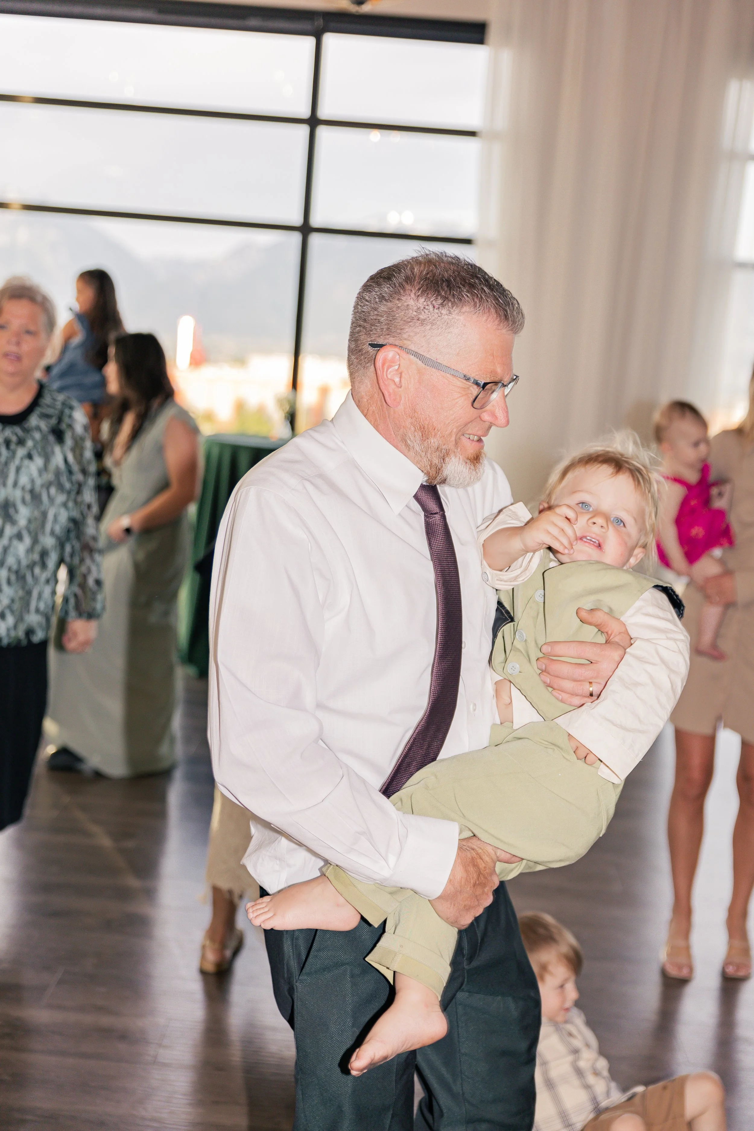 A man with glasses and a beard holds a young boy with blond hair and blue eyes in his arms during a social gathering indoors near a large window with a city view.