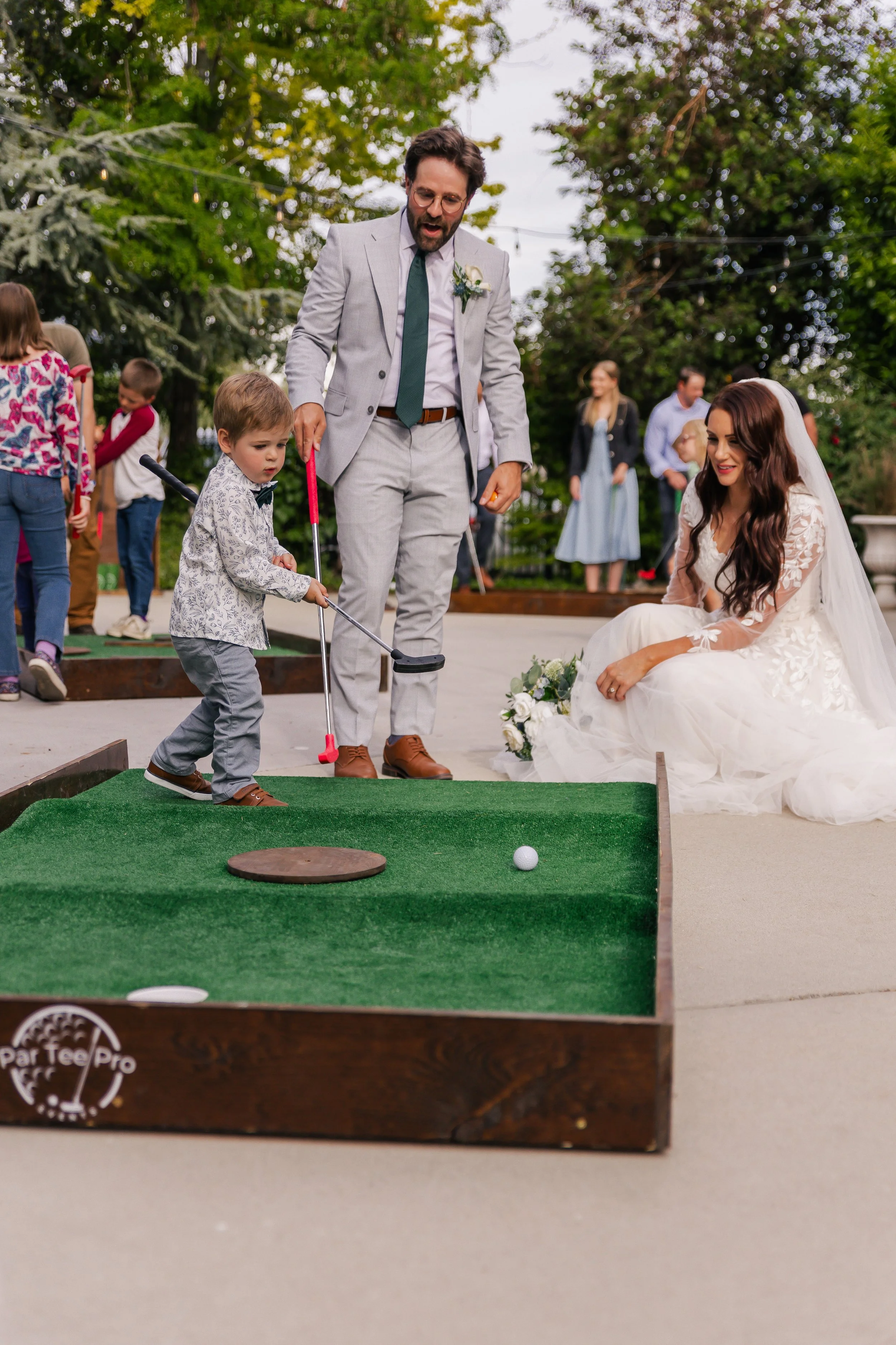 A bride and groom are celebrating their wedding outdoors, with children playing mini golf. A young boy is about to hit a golf ball while the bride smiles on, seated next to a bouquet of flowers. Guests are in the background, and trees are above.