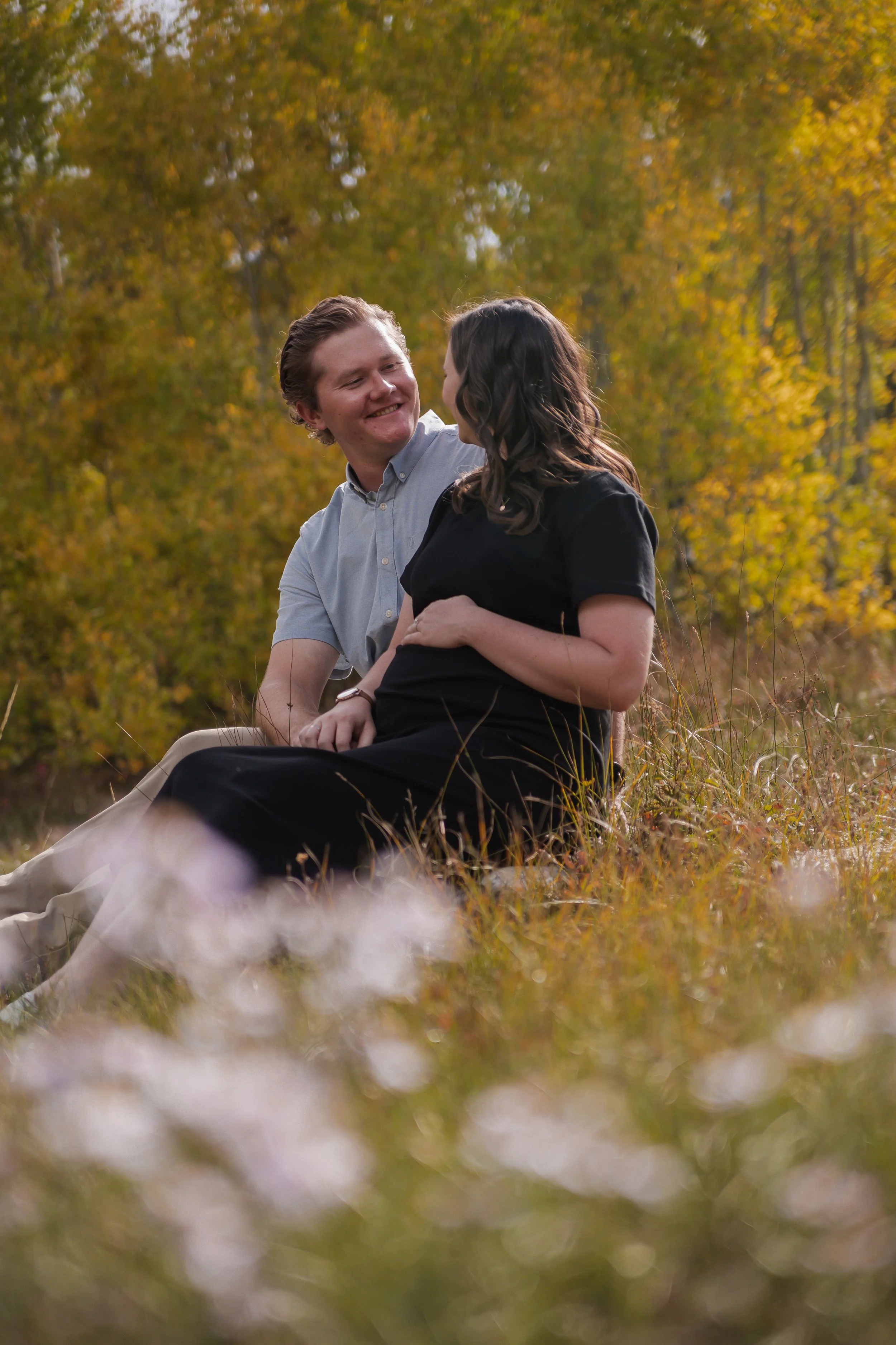 A couple sitting on grass in a park during autumn, smiling at each other.