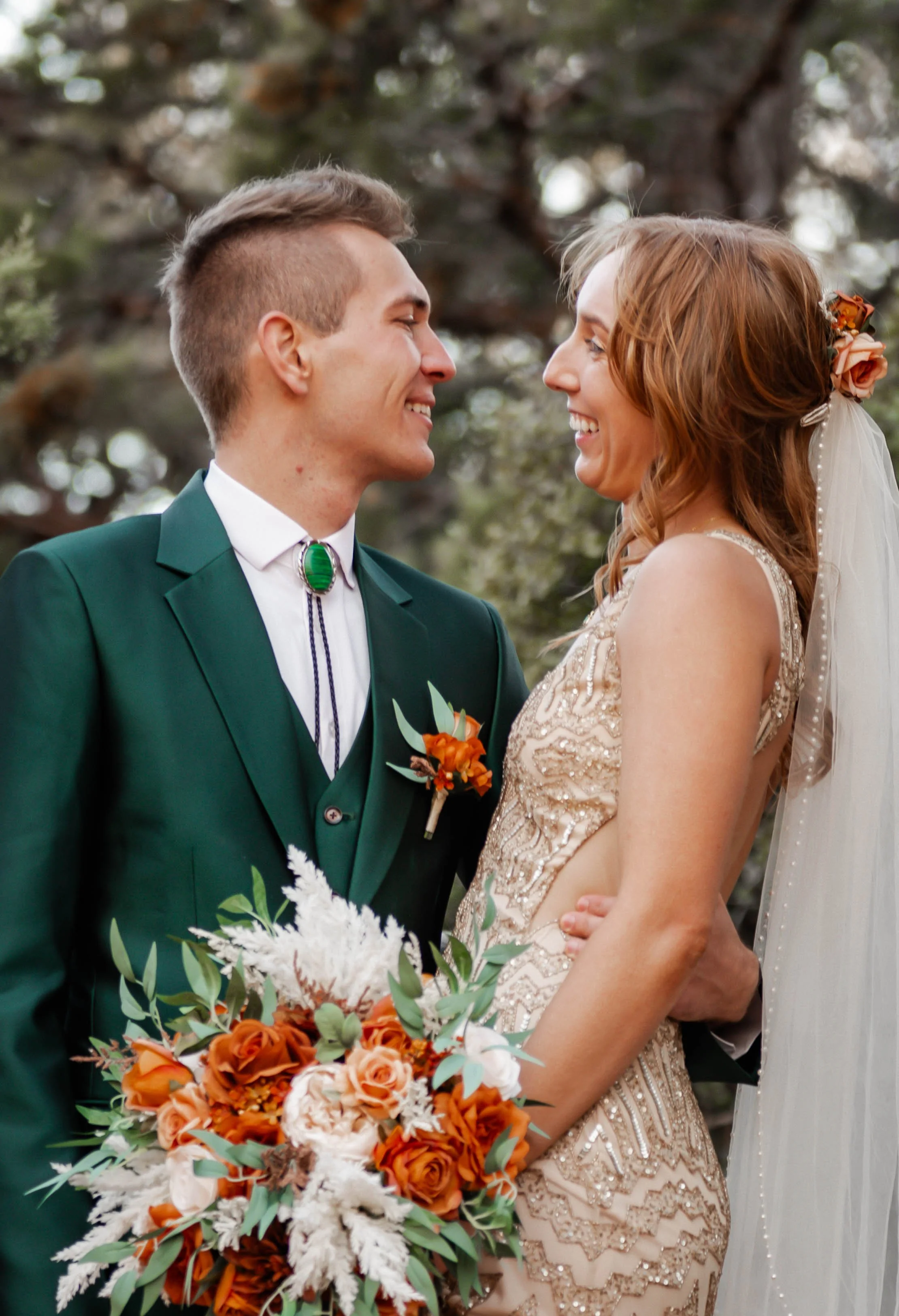 A bride and groom face each other smiling outdoors with trees in the background. The groom is wearing a green suit with a bolo tie and boutonniere, holding a bouquet of orange and white flowers. The bride is wearing a lace wedding dress with a veil a