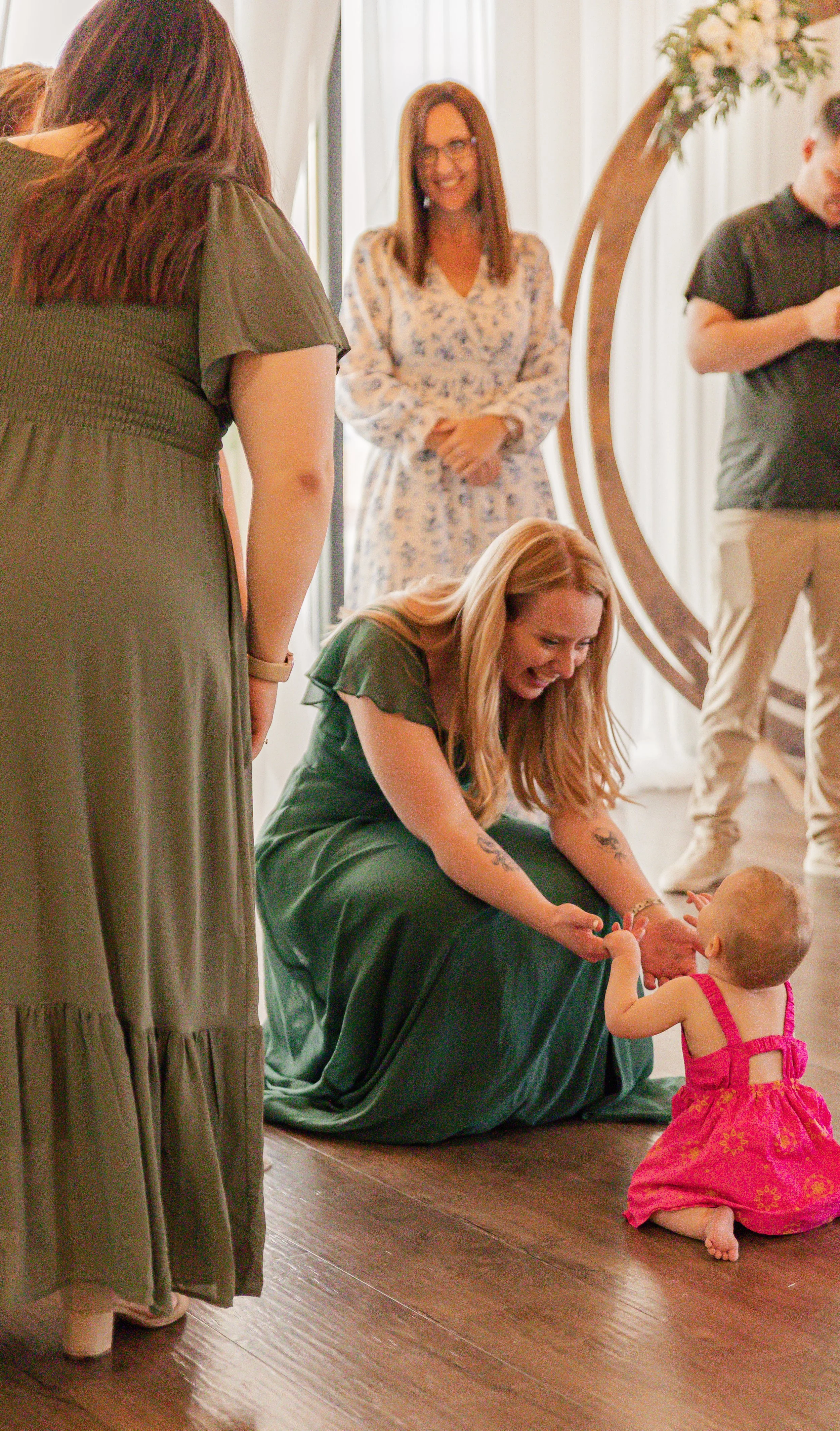 A woman in a green dress kneeling on the floor, playing with a young girl in a pink dress. Other women are standing and watching in the background, near a circle-shaped wooden decoration and a floral arrangement.