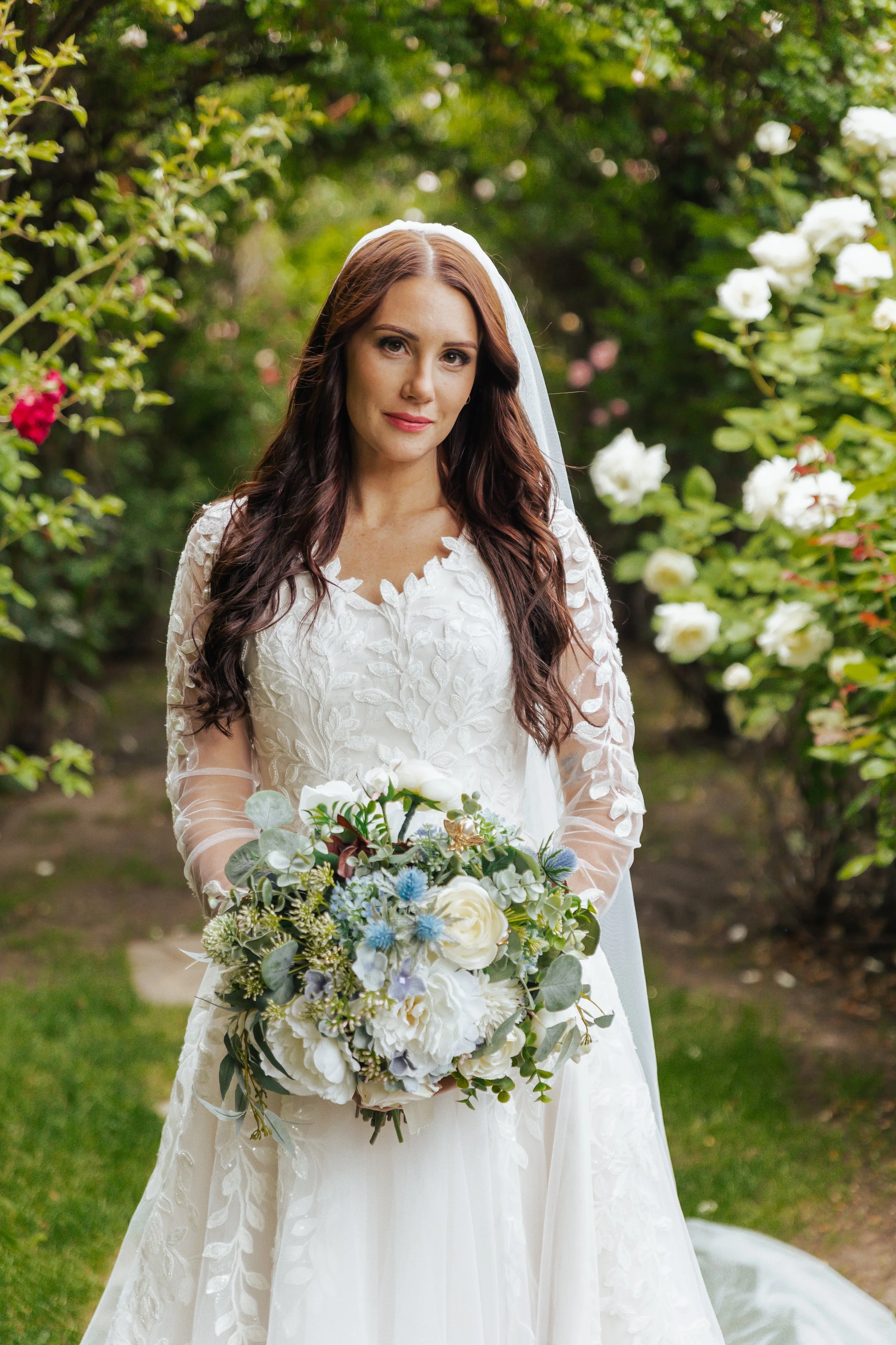 A bride standing outdoors in a garden, holding a bouquet of white and pastel flowers, wearing a wedding dress with lace details, and a veil, with greenery and white flowers in the background.