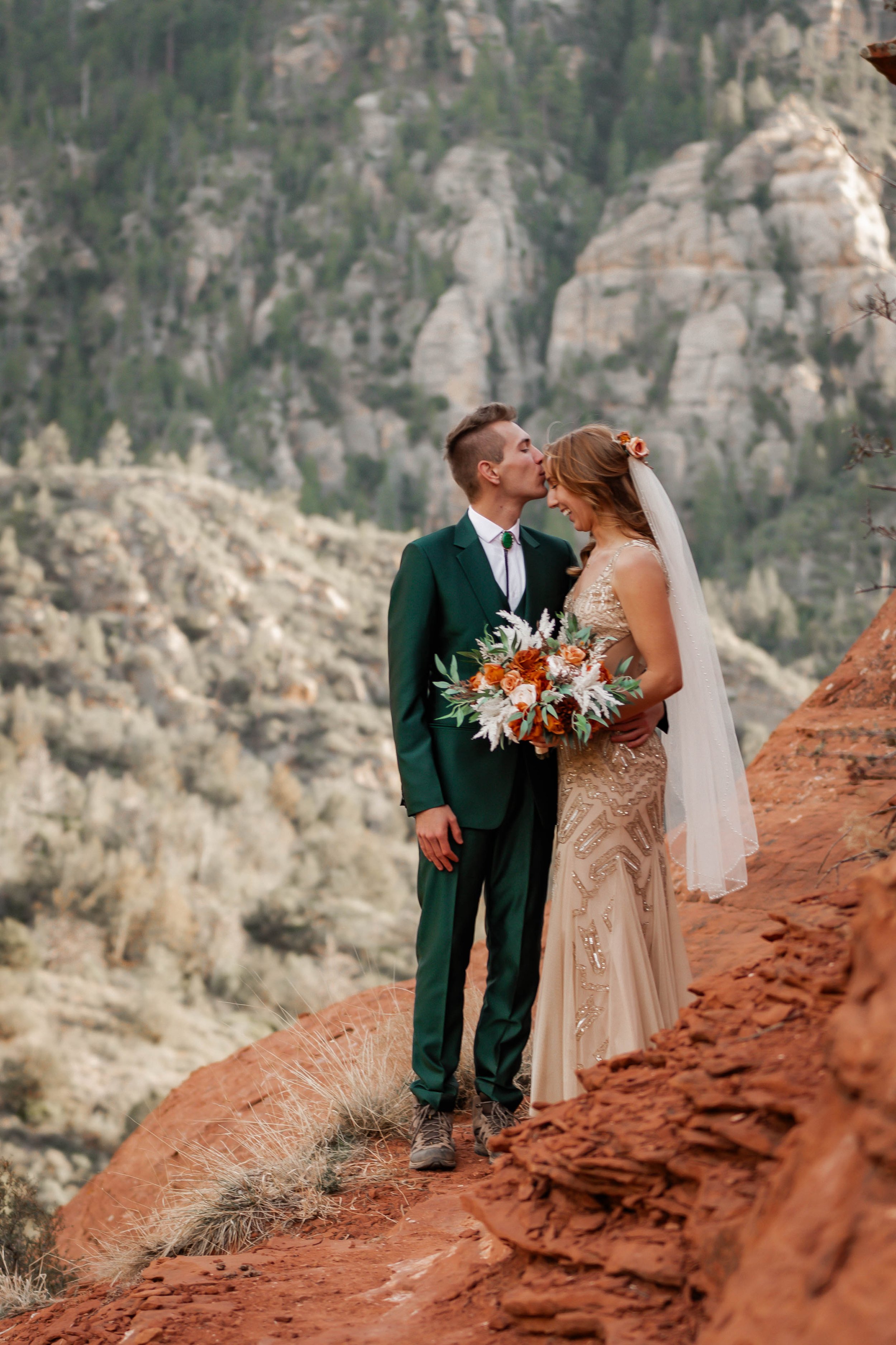 A newlywed couple standing on a rocky ledge in a mountainous landscape. The groom, dressed in a green suit, is kissing the bride's forehead. The bride is wearing a beige, beaded wedding gown and holding a bouquet of flowers, with her hair adorned wit