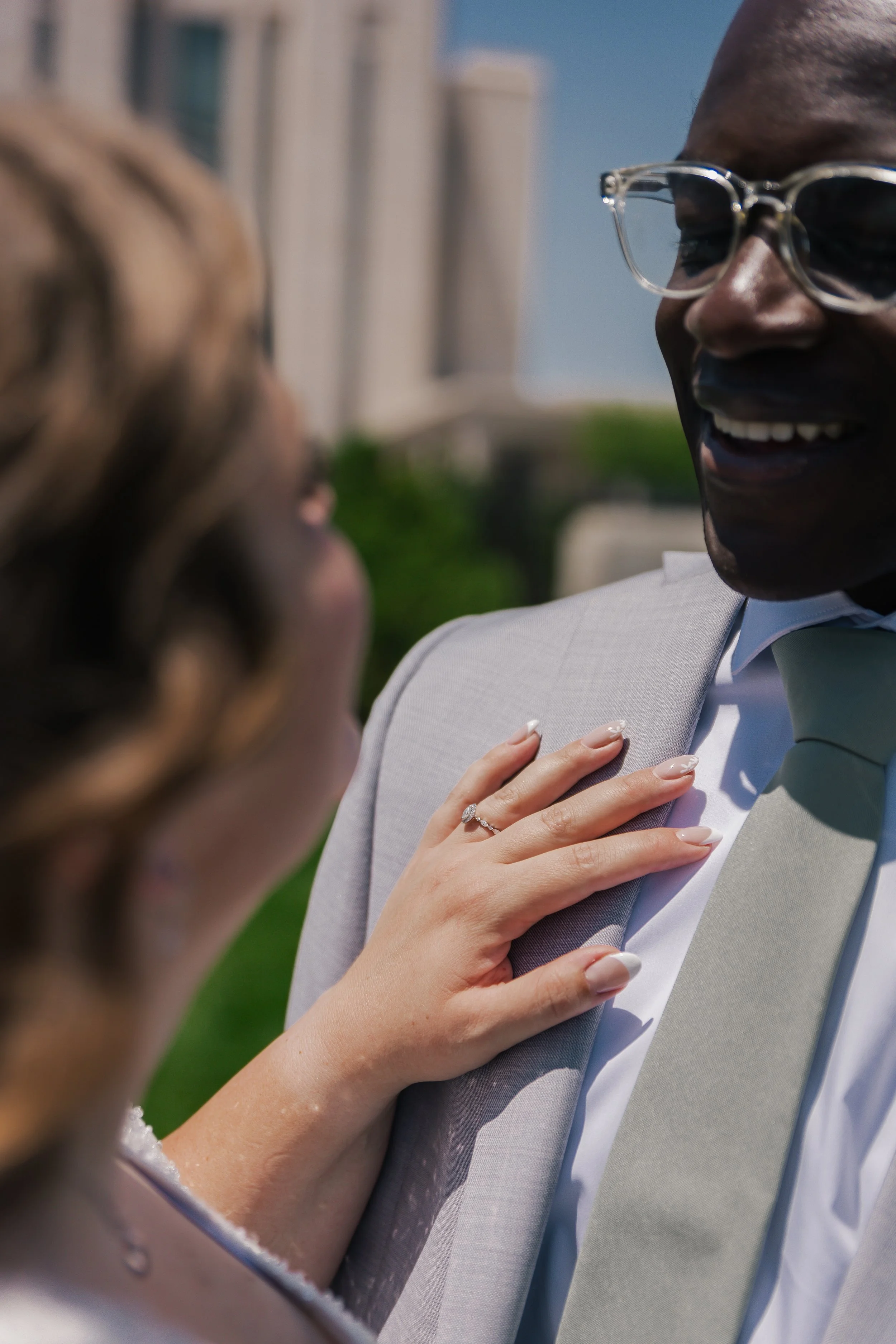 Close-up of a bride with a diamond ring touching the chest of a groom wearing glasses and a light gray suit during a wedding ceremony outdoors.