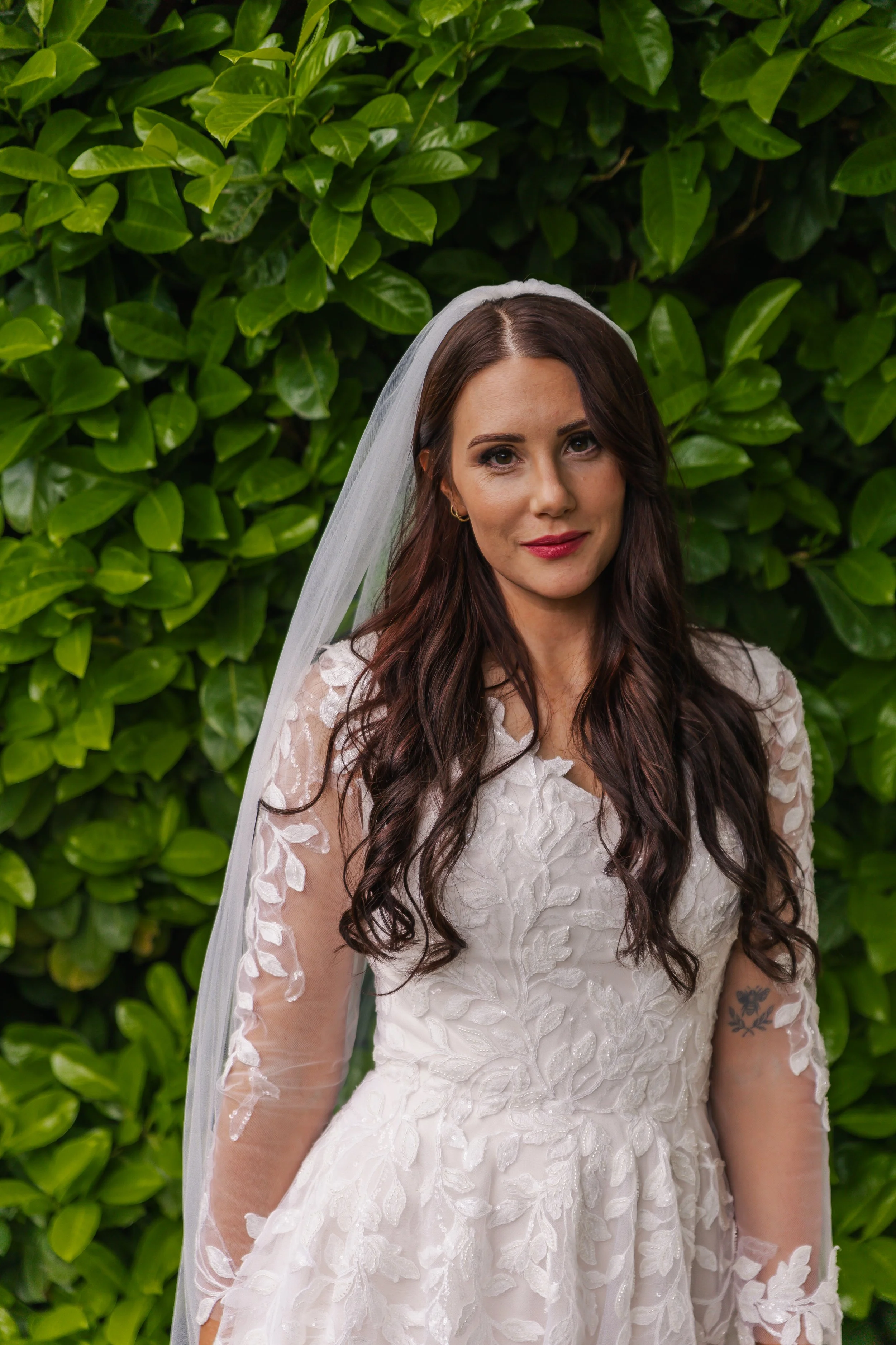 A woman in a white lace wedding dress with sheer sleeves, standing outdoors in front of green bushes, wearing a veil and makeup.