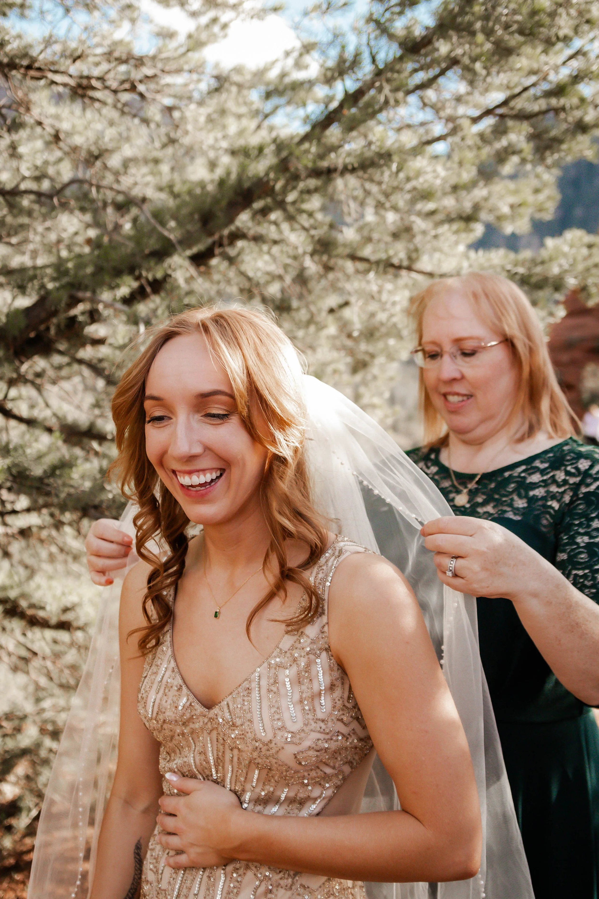 A bride with red hair wearing a beaded gown is smiling as an older woman helps her put on a veil outdoors during the daytime, with blooming trees in the background.
