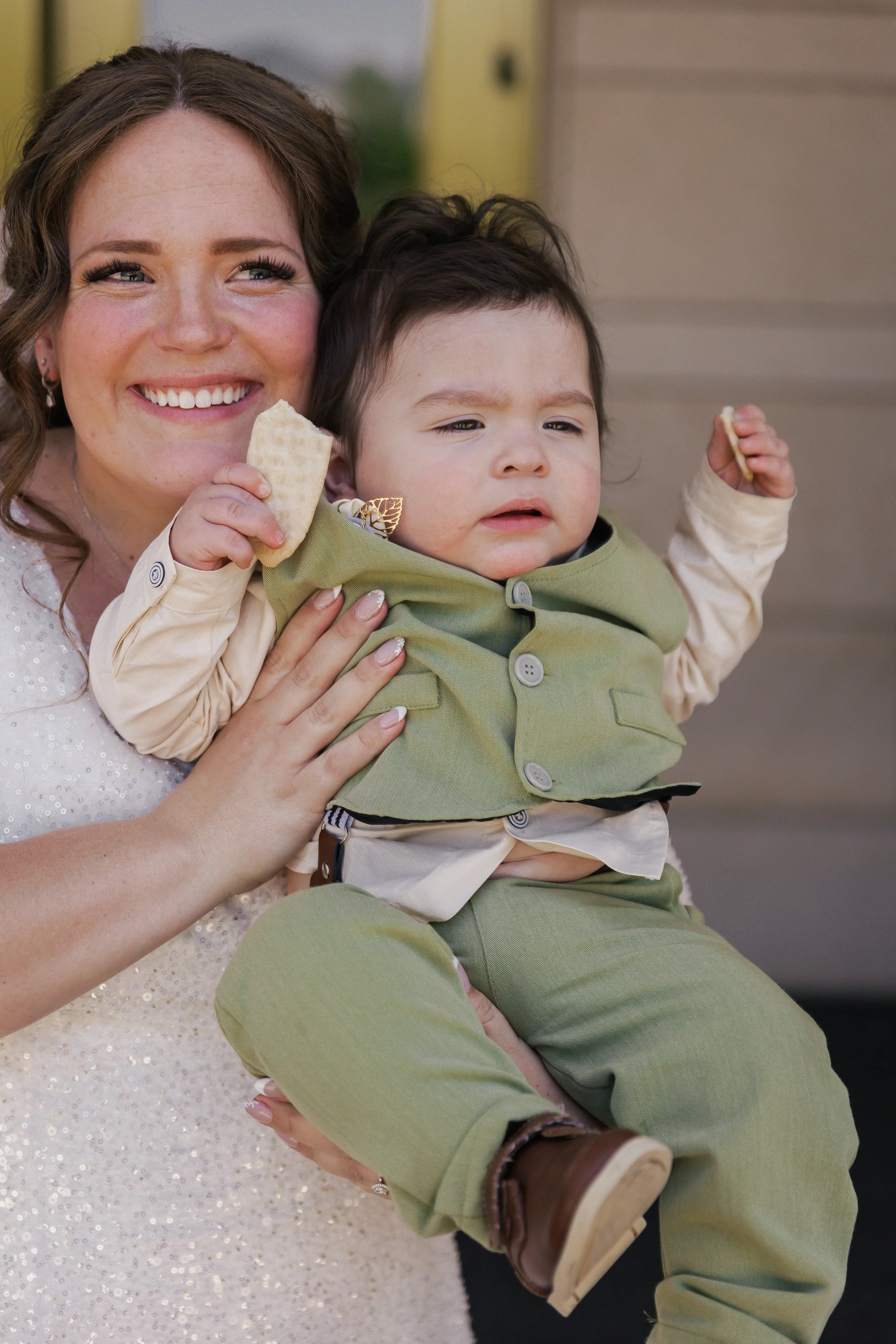 A woman with curly brown hair and wearing a white sparkly dress holds a young boy with dark hair, both smiling and holding waffles.