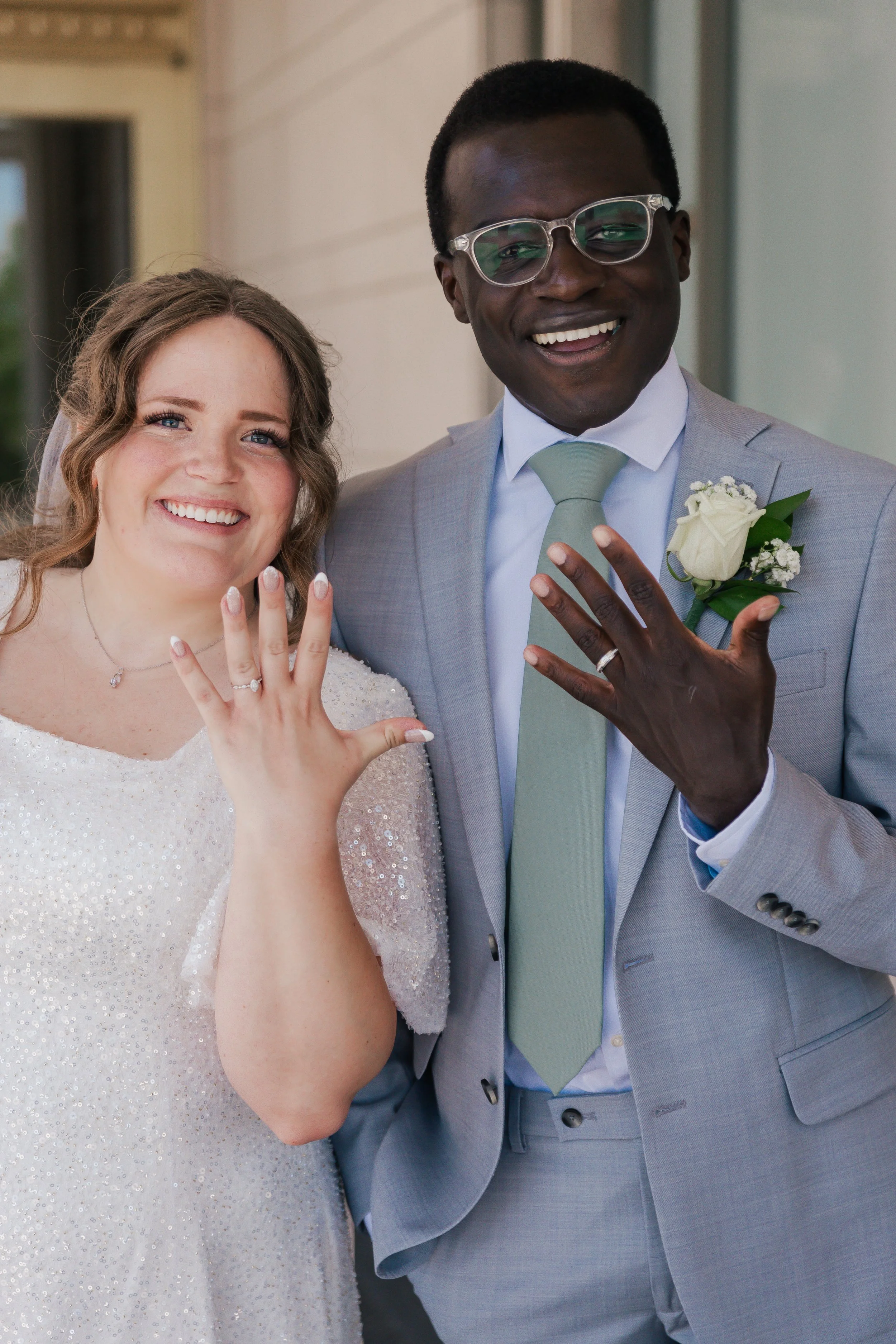 A happy wedding couple showing off their rings, standing outside a building. The bride is wearing a white, beaded dress and the groom is in a light gray suit with a green tie, glasses, and a white flower boutonniere.