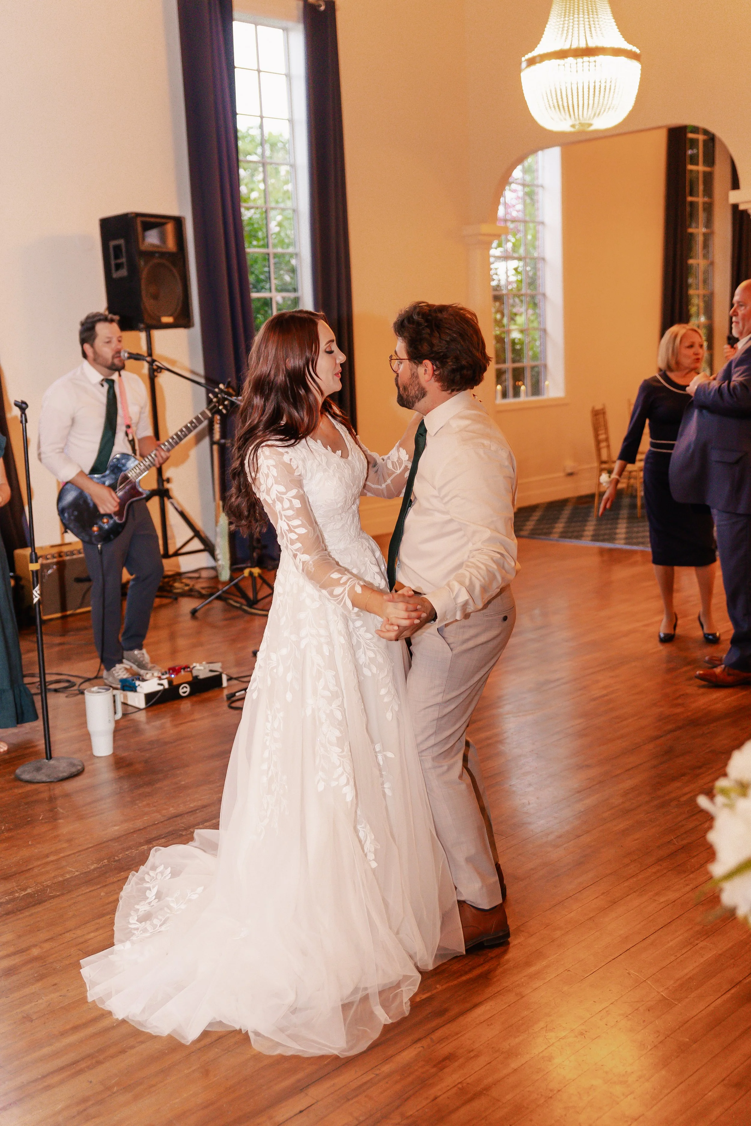 A bride and groom are dancing together at a wedding reception, with live band music playing in the background and other guests socializing.