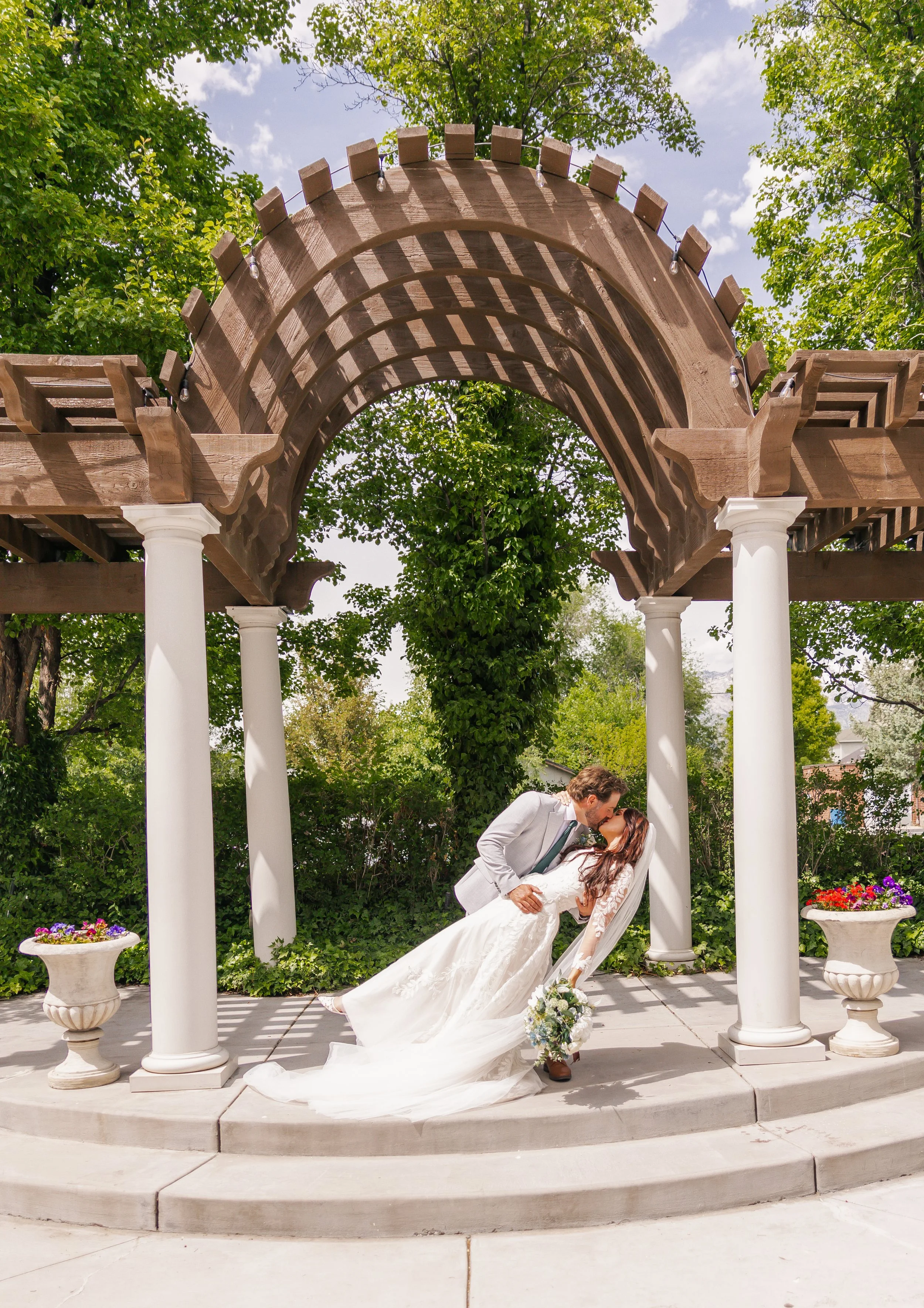 A bride and groom sharing a kiss under a wooden arbor with white columns, surrounded by trees and flower pots.
