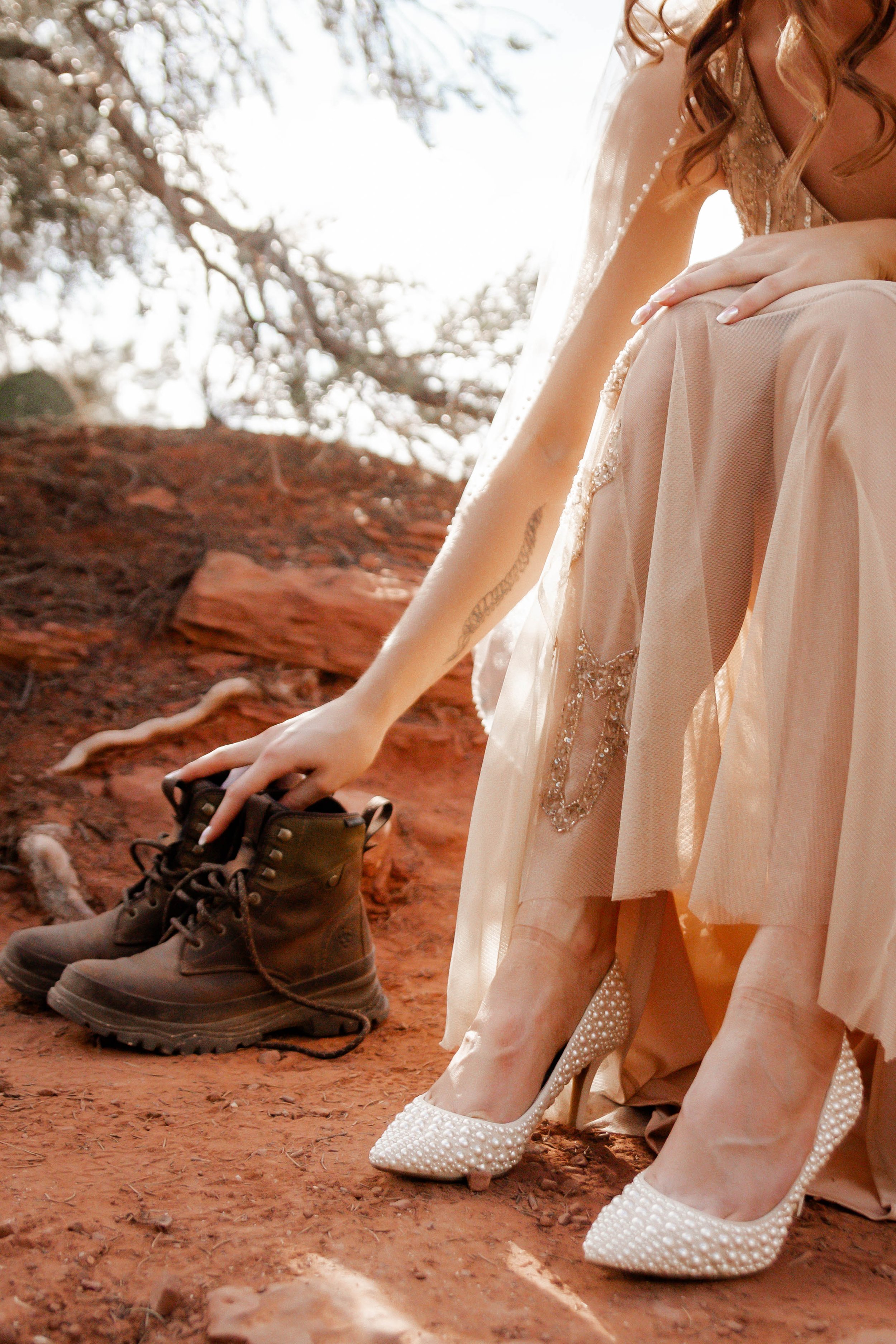 A woman in wedding attire, wearing white high heels with pearl embellishments, is reaching down to pick up a pair of hiking boots on a dirt ground outdoors.