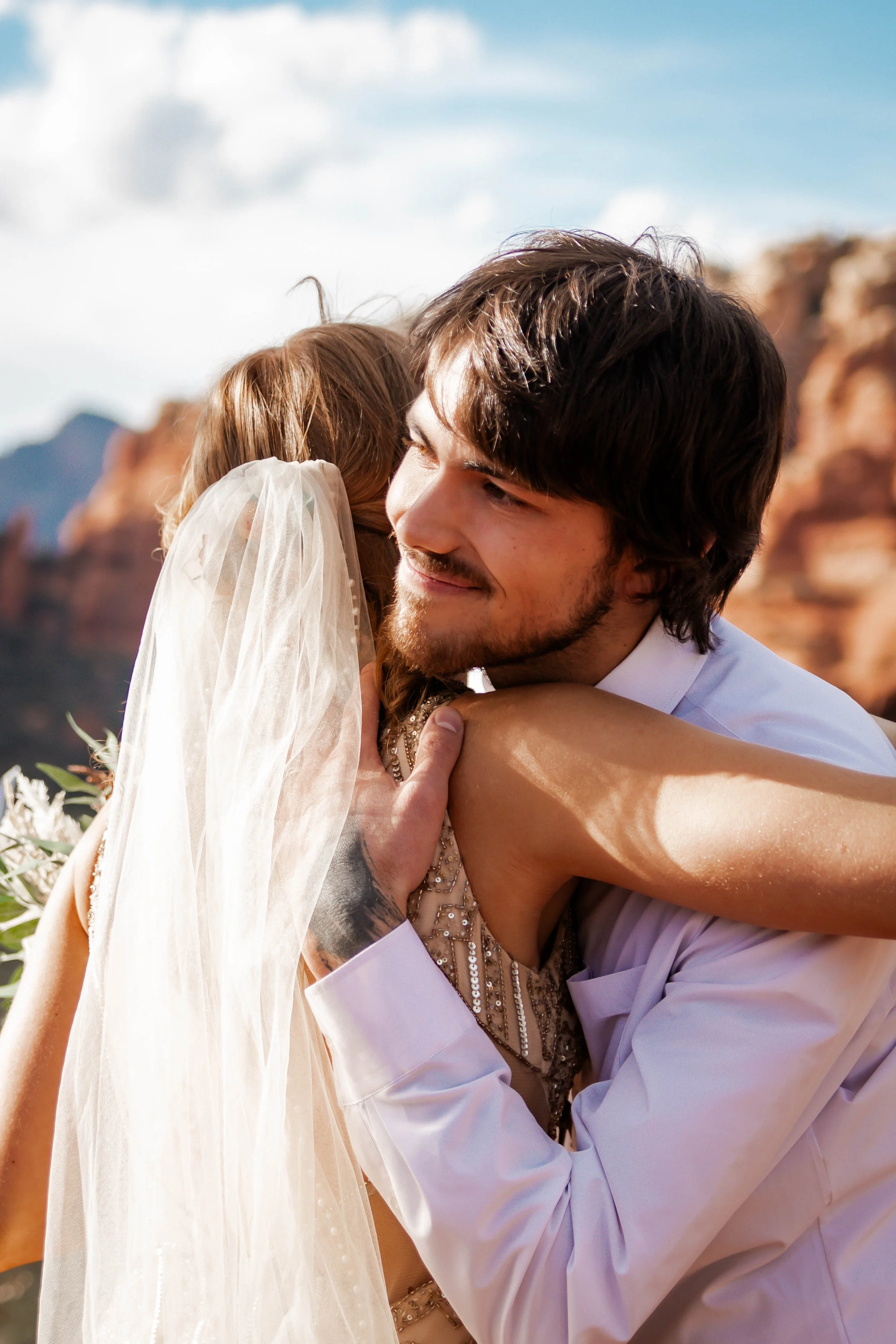 A bride and groom sharing a warm hug outdoors with a rocky, desert landscape in the background.