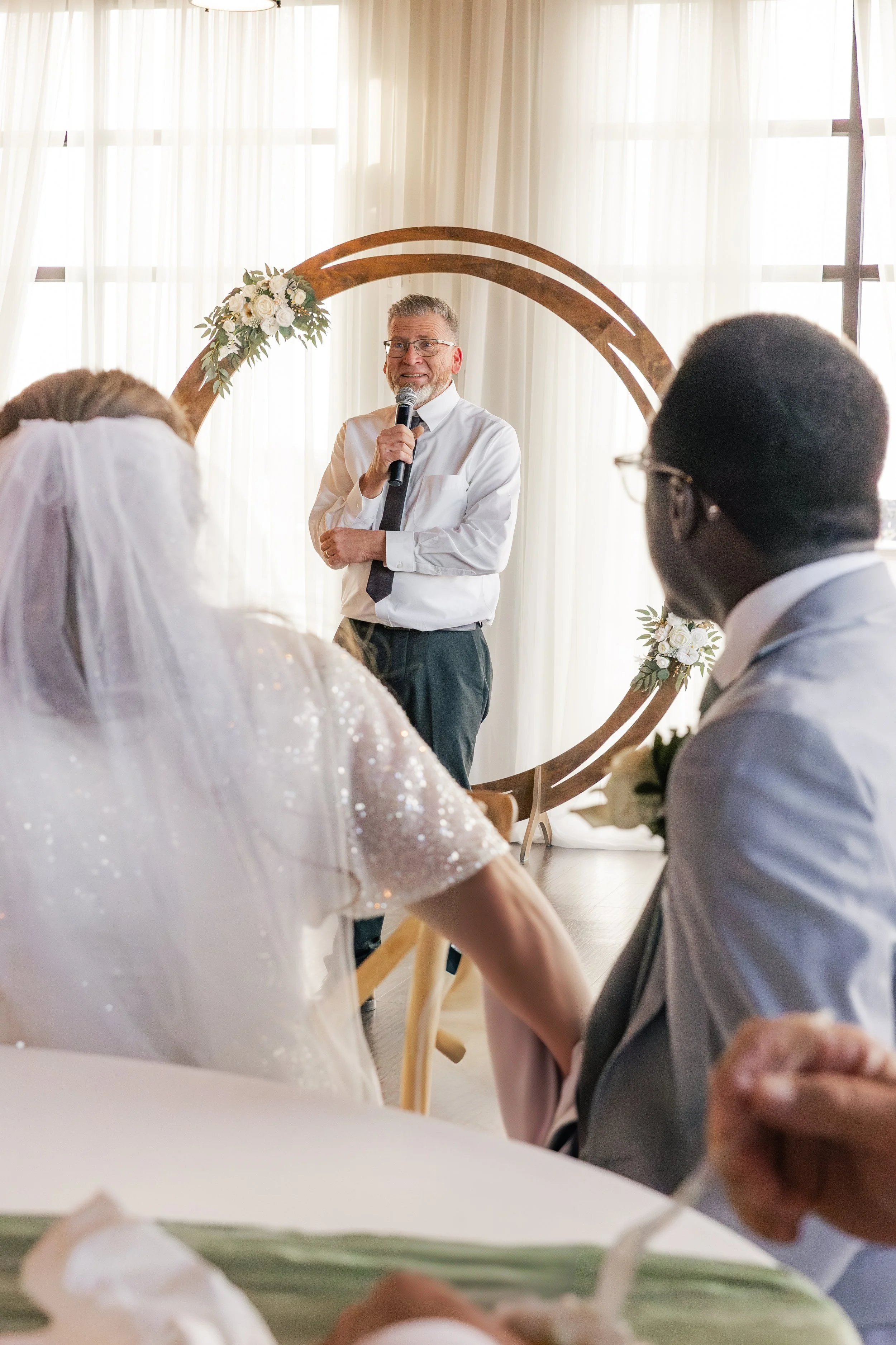 A man giving a speech at a wedding, standing in front of a decorative floral arch, with guests listening attentively.