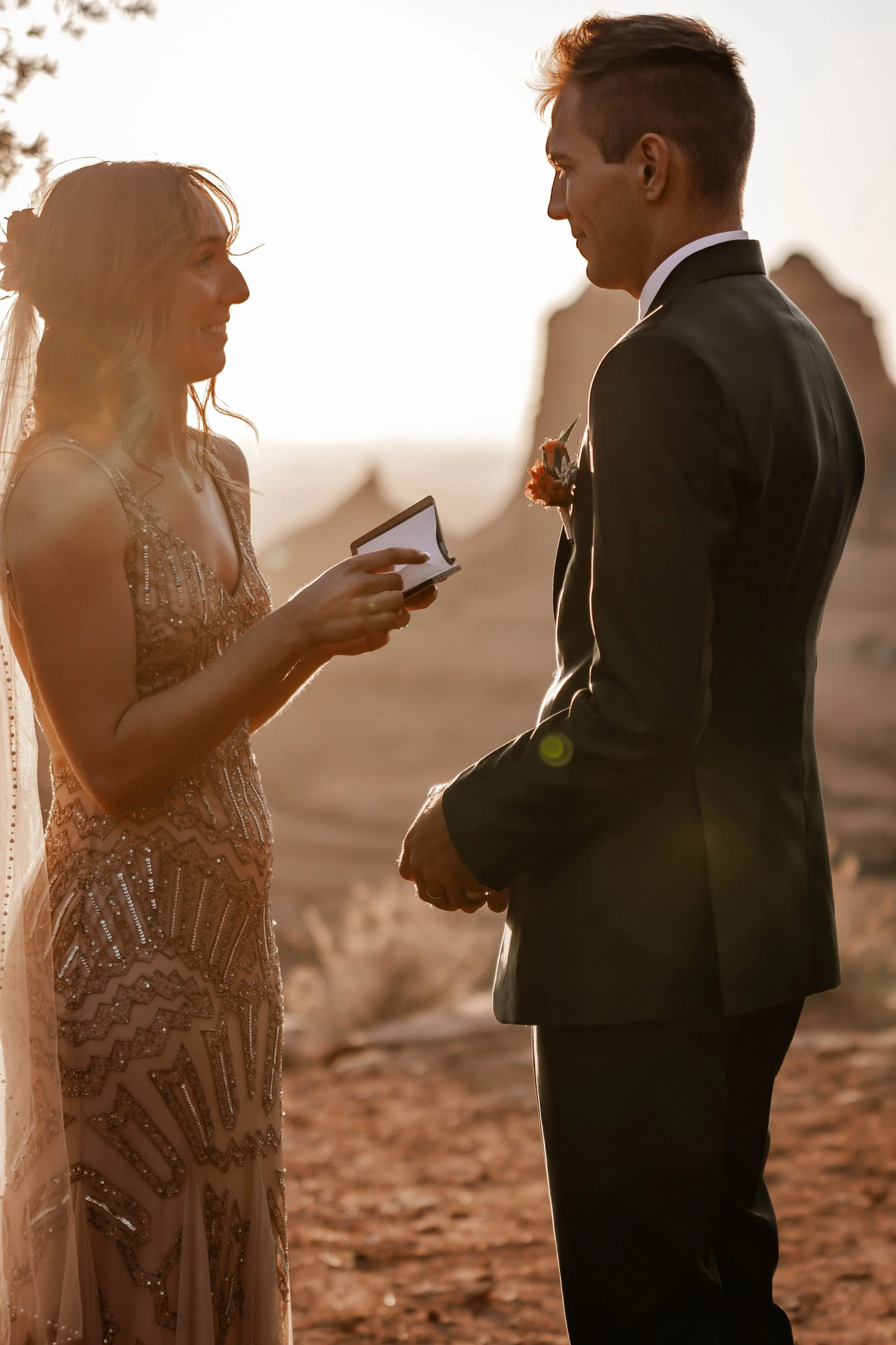 A couple getting married outdoors during sunset, with the bride holding a small book or paper and the groom in a black tuxedo, holding hands, in a desert-like setting with rock formations in the background.