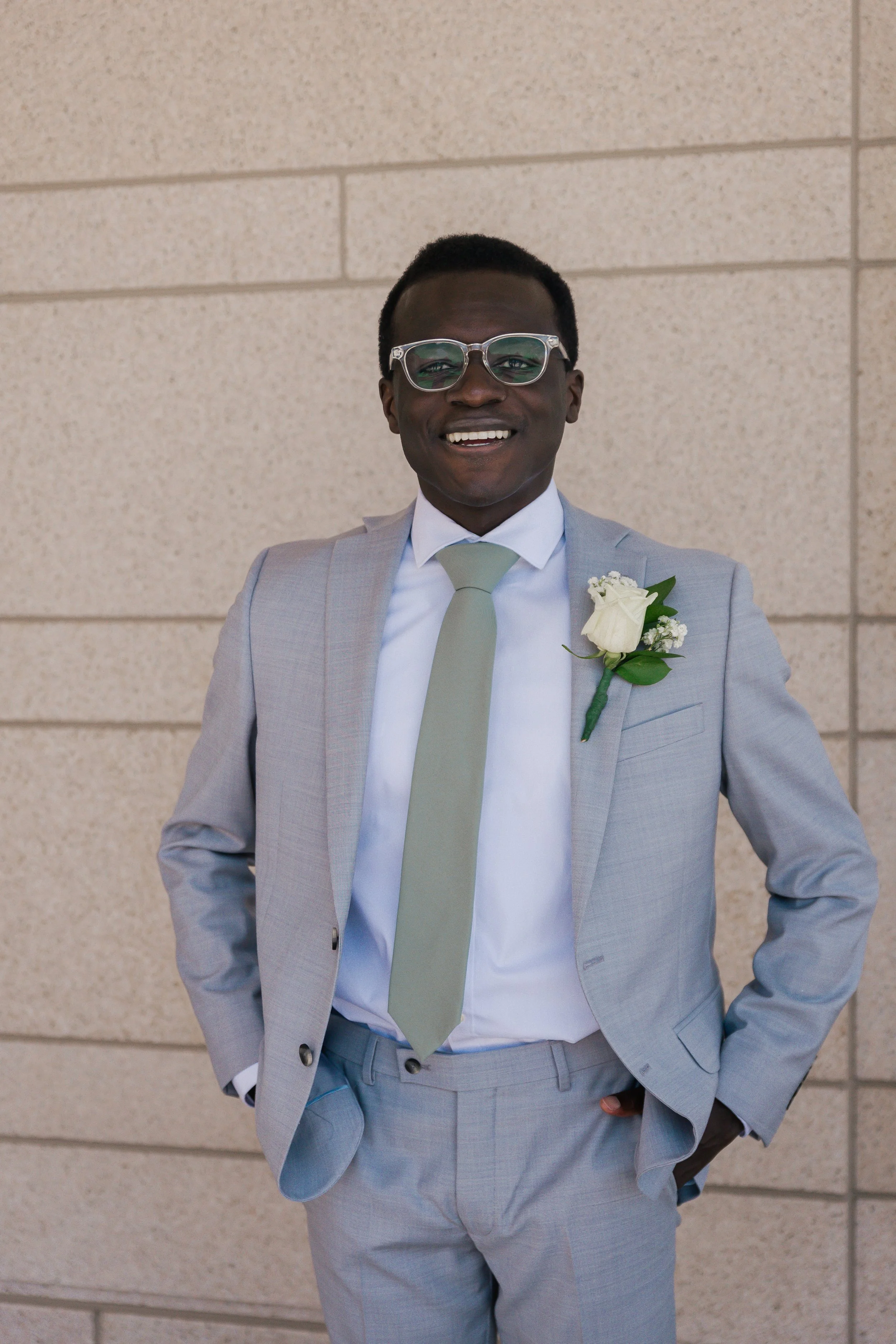 A man in a light gray suit with a matching tie and white shirt, smiling, standing against a beige brick wall. He is wearing glasses and has a white boutonniere on his lapel.