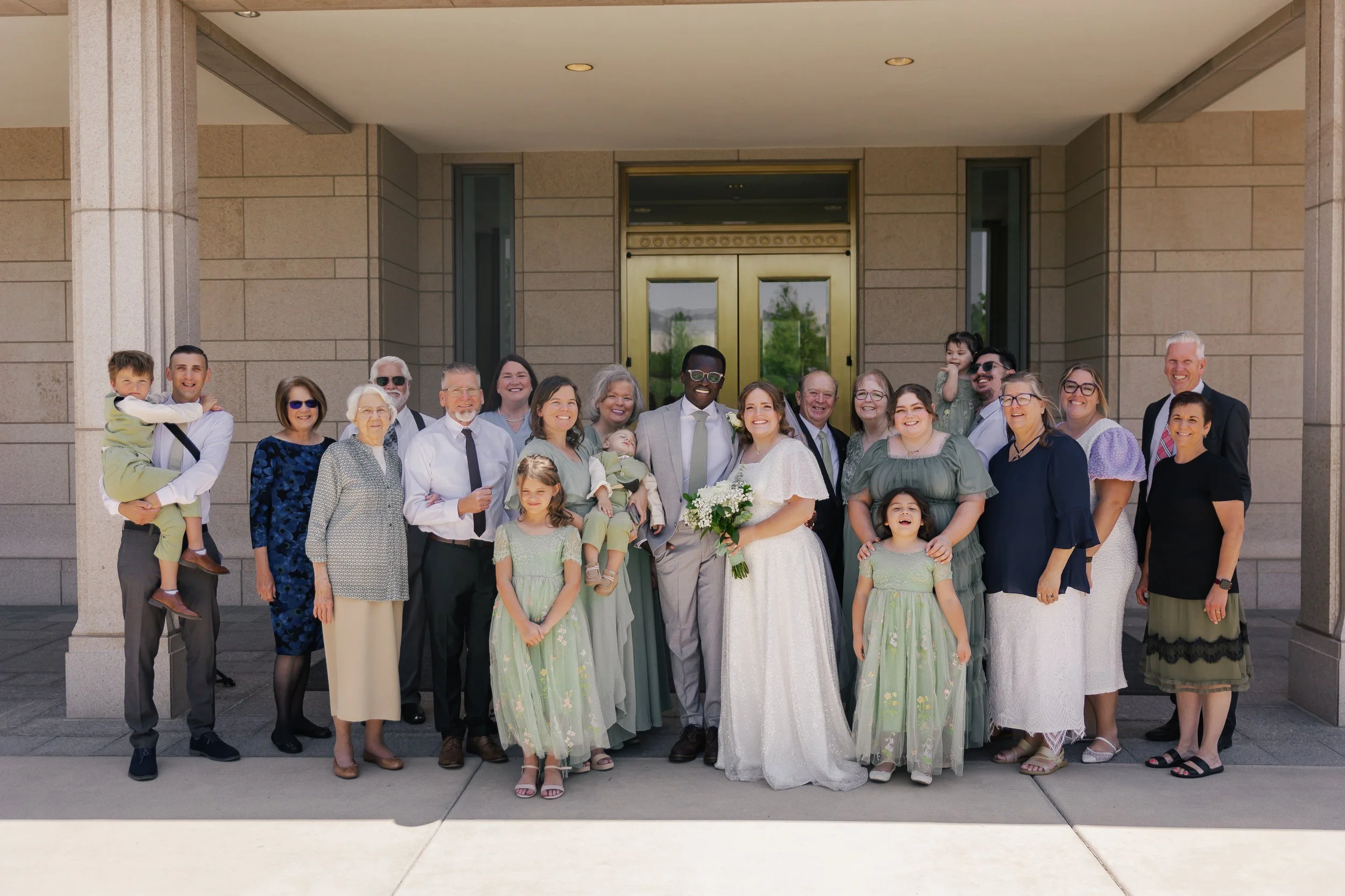 A large group of people, including a newlywed couple, stands in front of a building entrance for a wedding photo. The bride and groom are in the center, smiling, surrounded by family and friends.