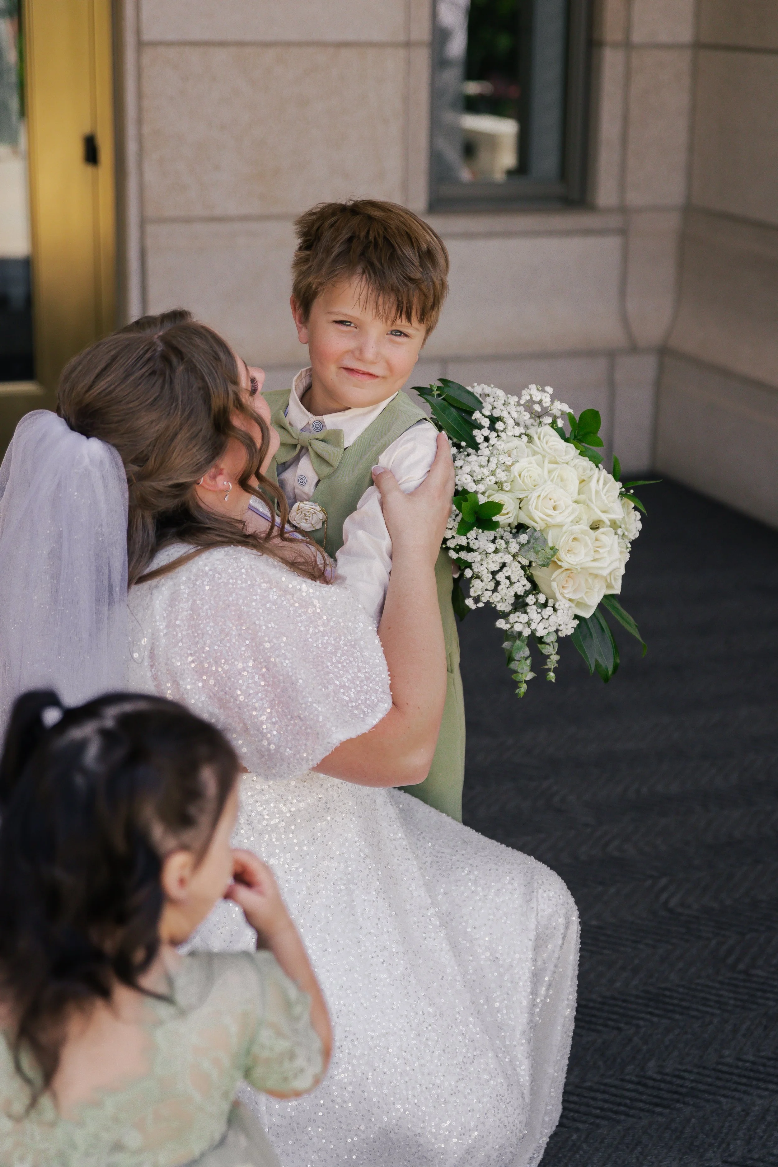 A woman in a white wedding dress holding a young boy with a bouquet of white roses, in an indoor setting, with two young girls nearby.