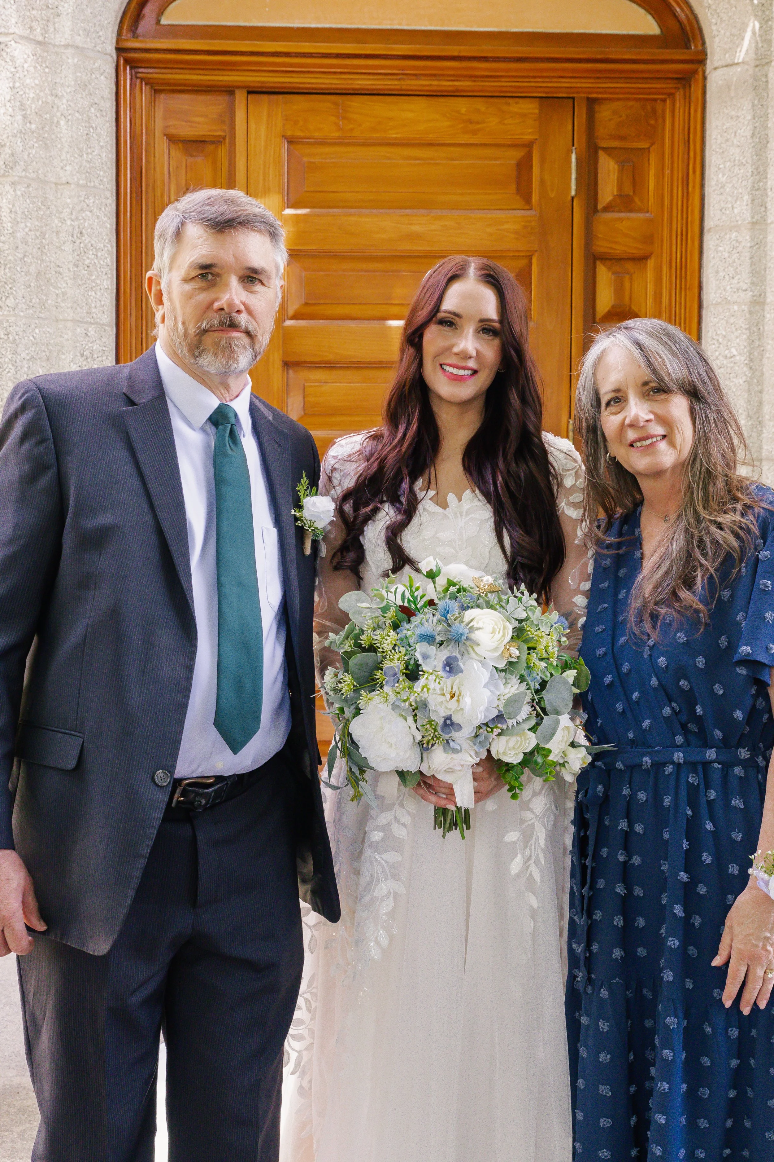 A bride in a wedding dress holding a bouquet of white, blue, and green flowers, standing between a man in a suit and a woman in a blue dress, in front of a wooden door.