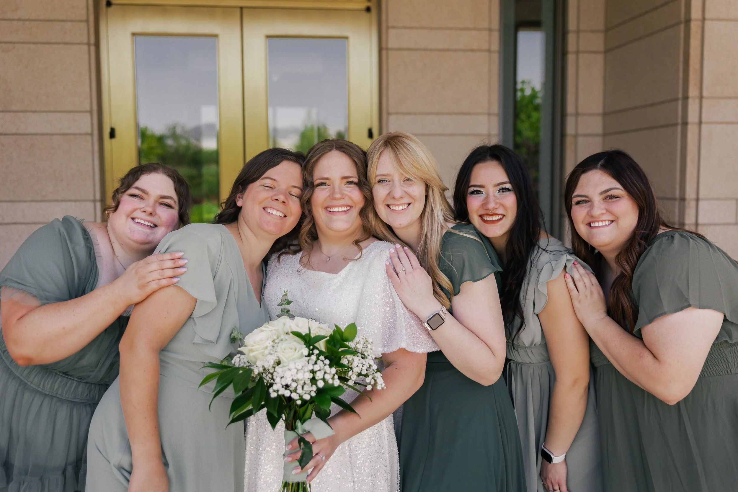 Group of six women smiling outside a building, with one woman in a white dress holding a bouquet, likely a bride, surrounded by five women in matching green dresses, possibly bridesmaids.