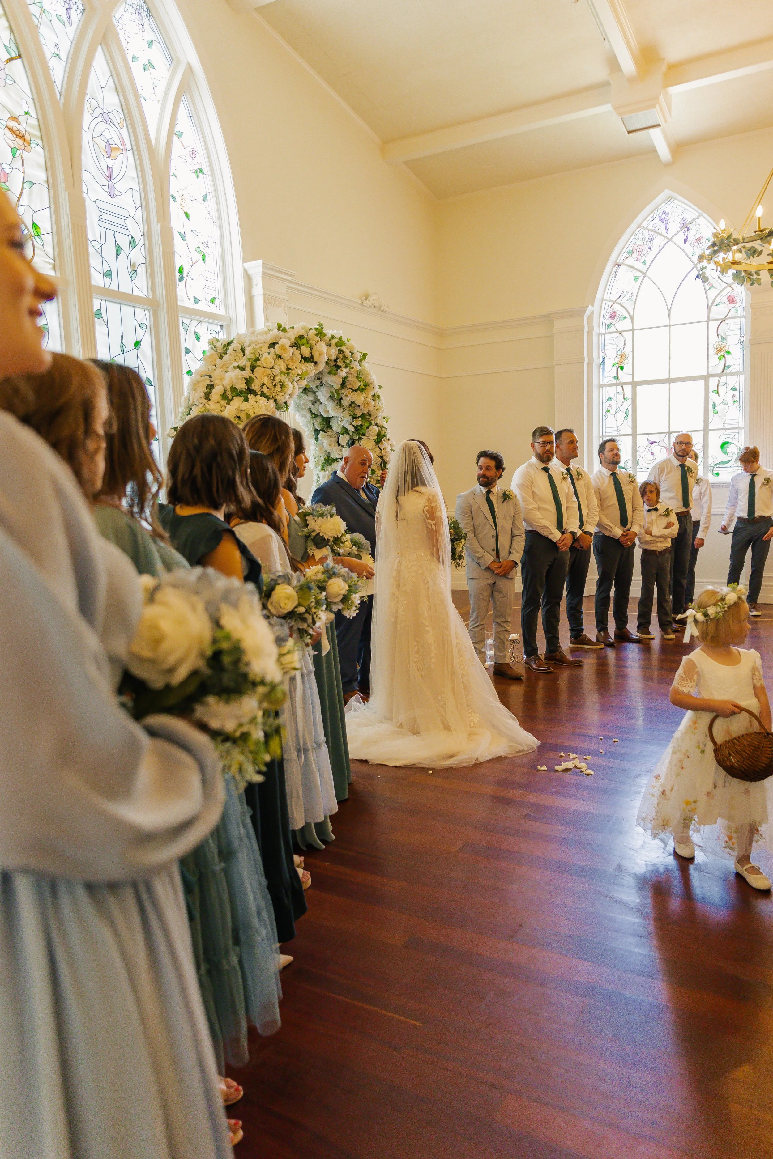 A wedding ceremony taking place inside a church with stained glass windows, featuring the bride and groom at the altar surrounded by bridesmaids and groomsmen. A flower girl in a white dress with a floral crown is walking.