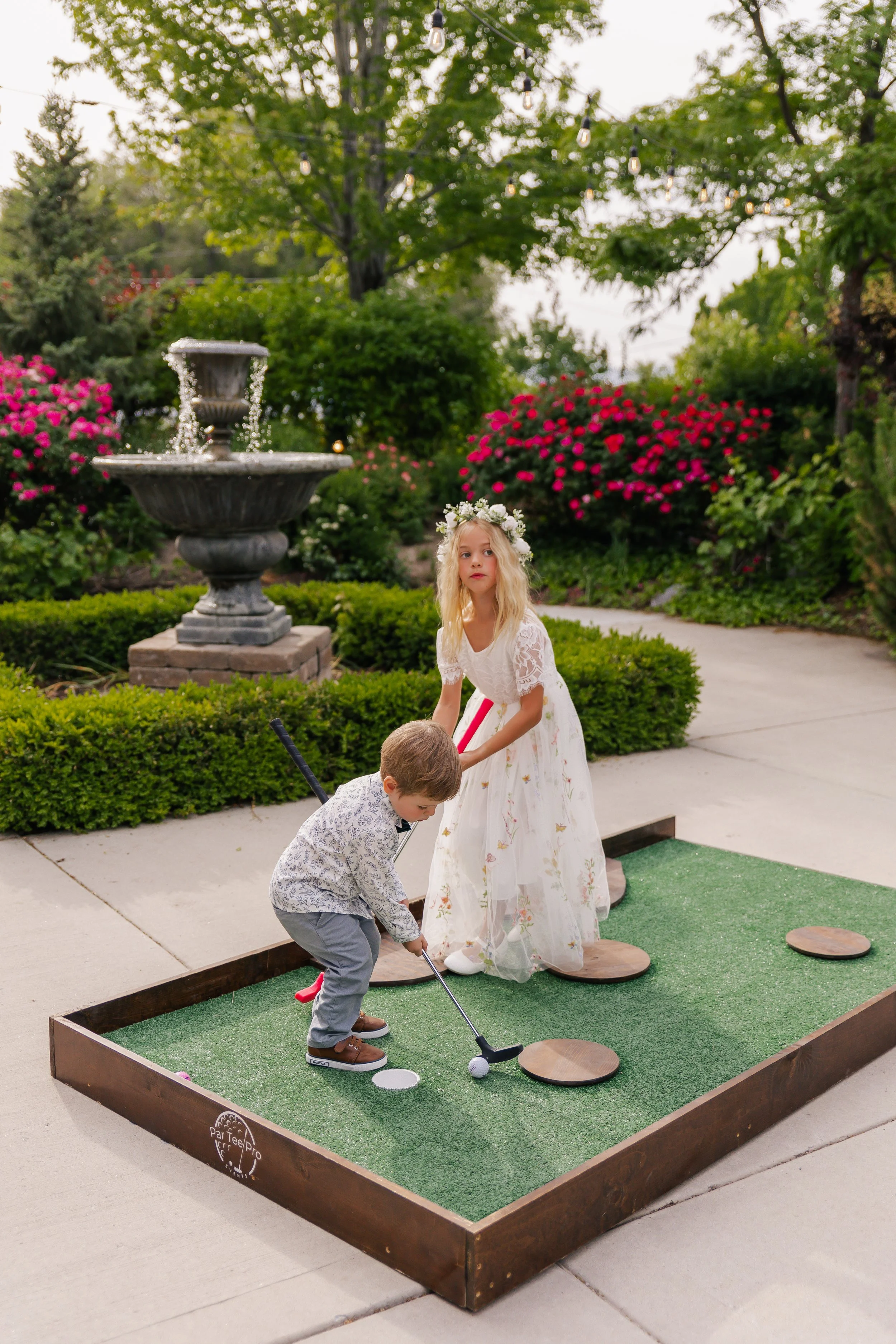 Two children playing mini golf on a green platform outdoors, surrounded by greenery and colorful flowers, with string lights overhead and a fountain in the background.