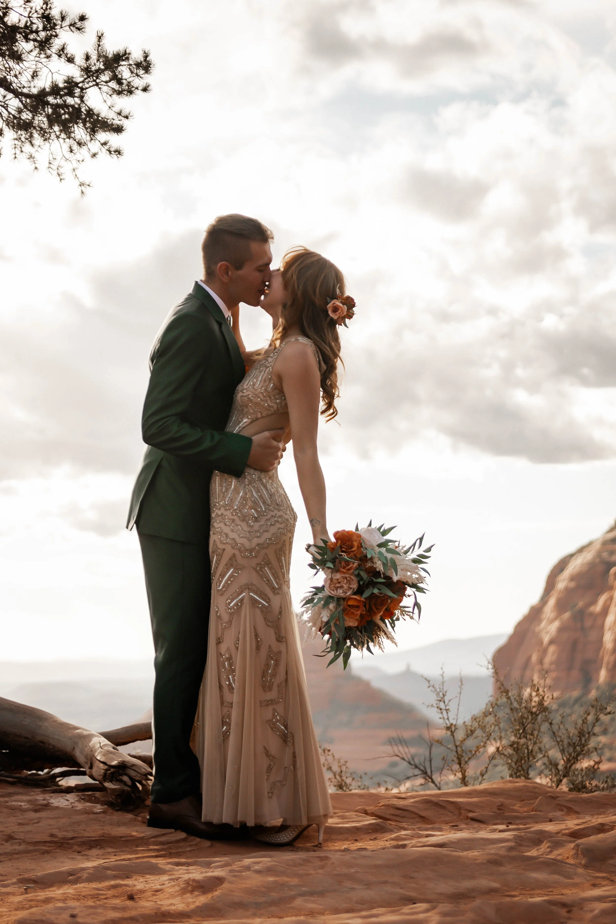 A bride and groom sharing a kiss outdoors during their wedding ceremony, with a scenic backdrop of mountains and cloudy sky, the bride holding a bouquet of flowers.