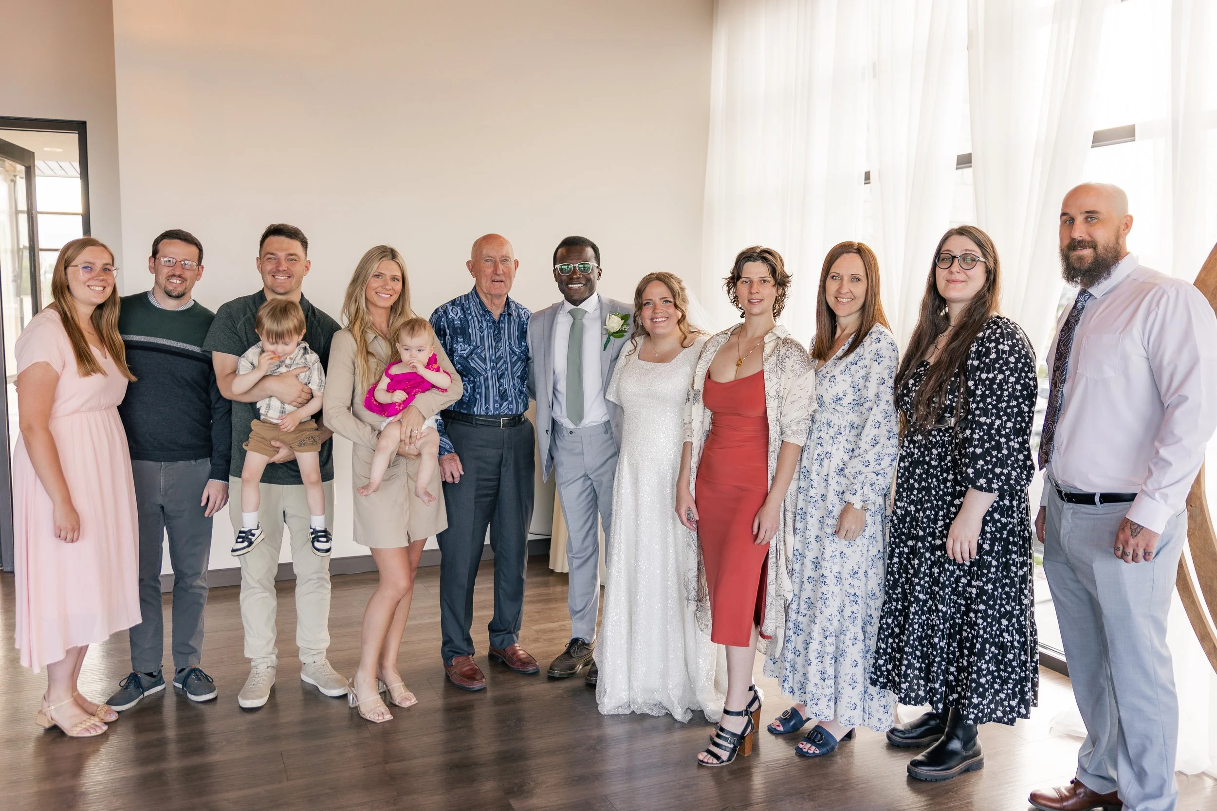 Group of people at a wedding celebration, including a bride and groom, family members, and friends, standing indoors in front of large windows with sheer curtains.