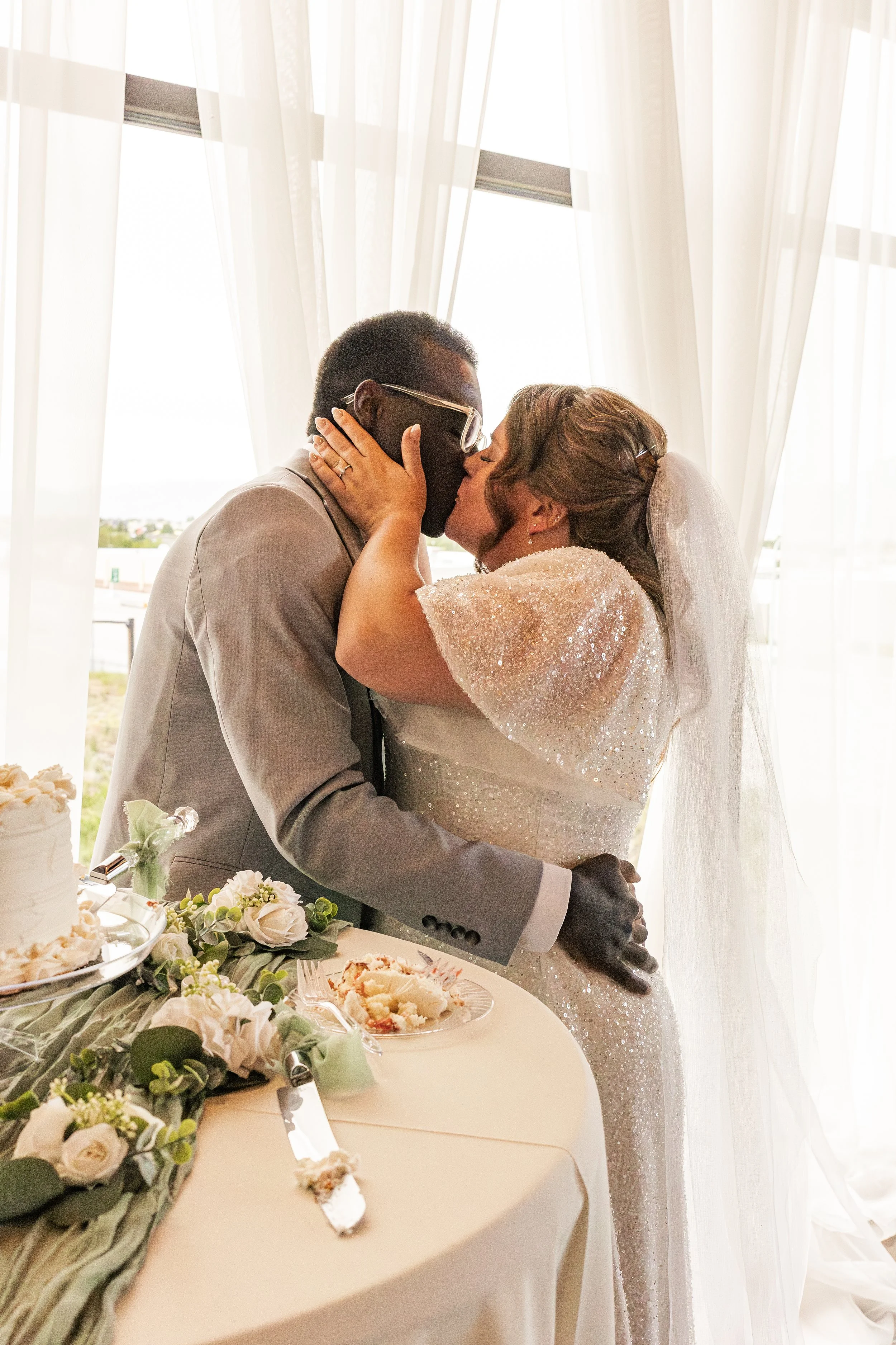 A same-sex couple shares a kiss at their wedding reception, with wedding cake and flowers on the table in front of them, and curtains with sunlight in the background.