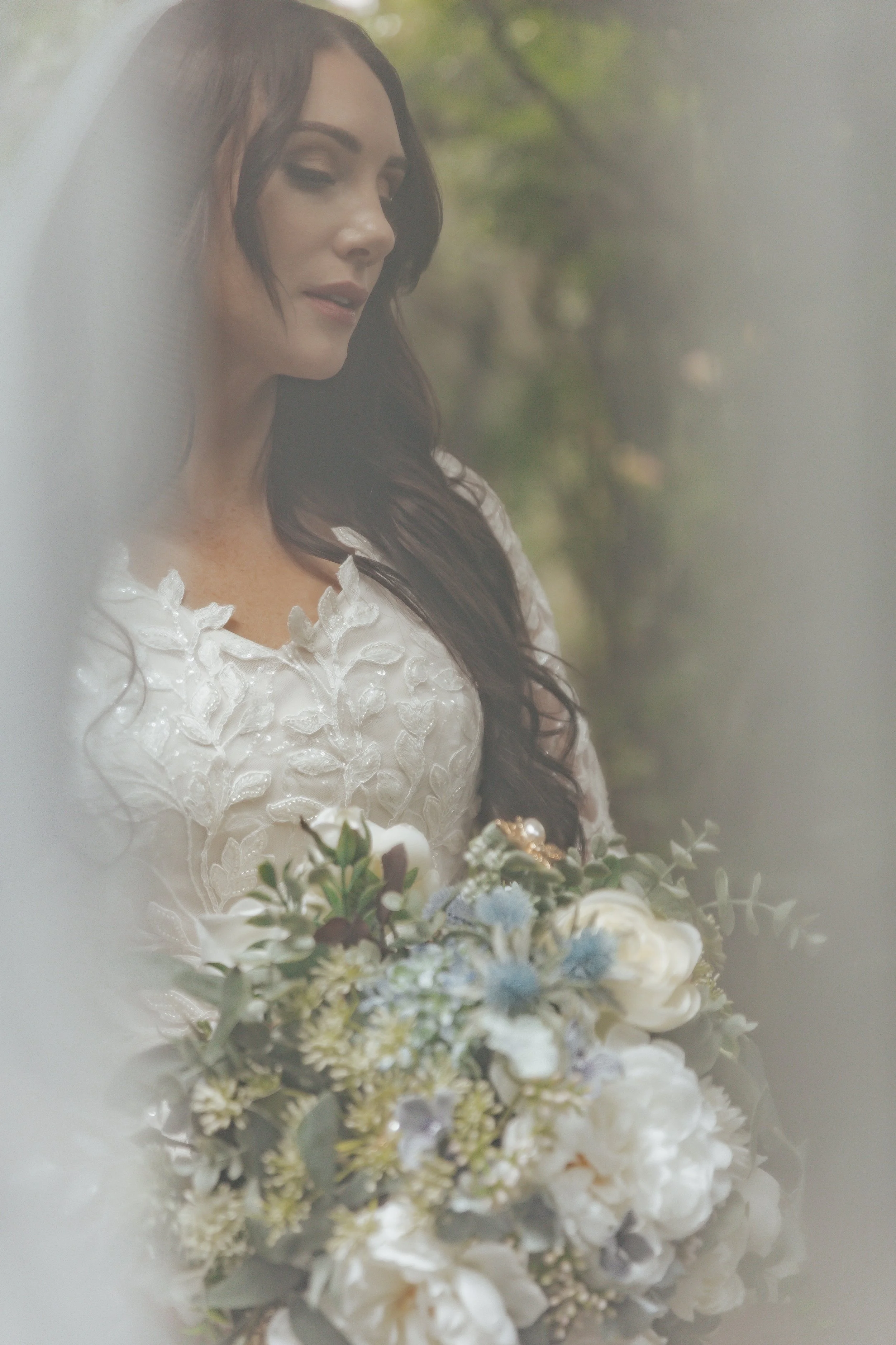 A bride with long dark hair wearing an embroidered white wedding dress holding a bouquet of white and blue flowers, standing outdoors.