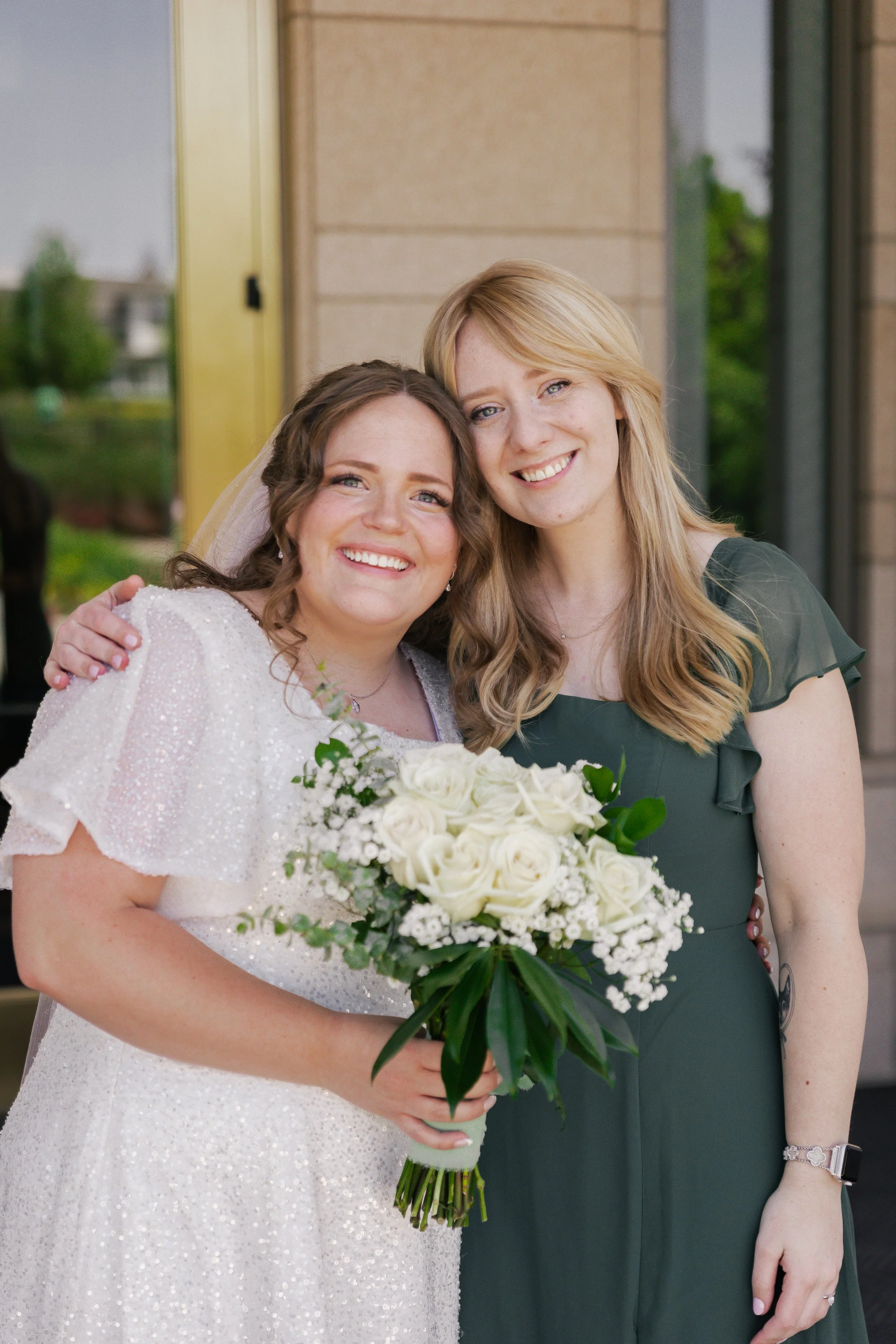 Two women, one dressed as a bride with a white, sparkly dress and veil, and the other in a green dress, smiling and holding a bouquet of white roses, standing outside a building.