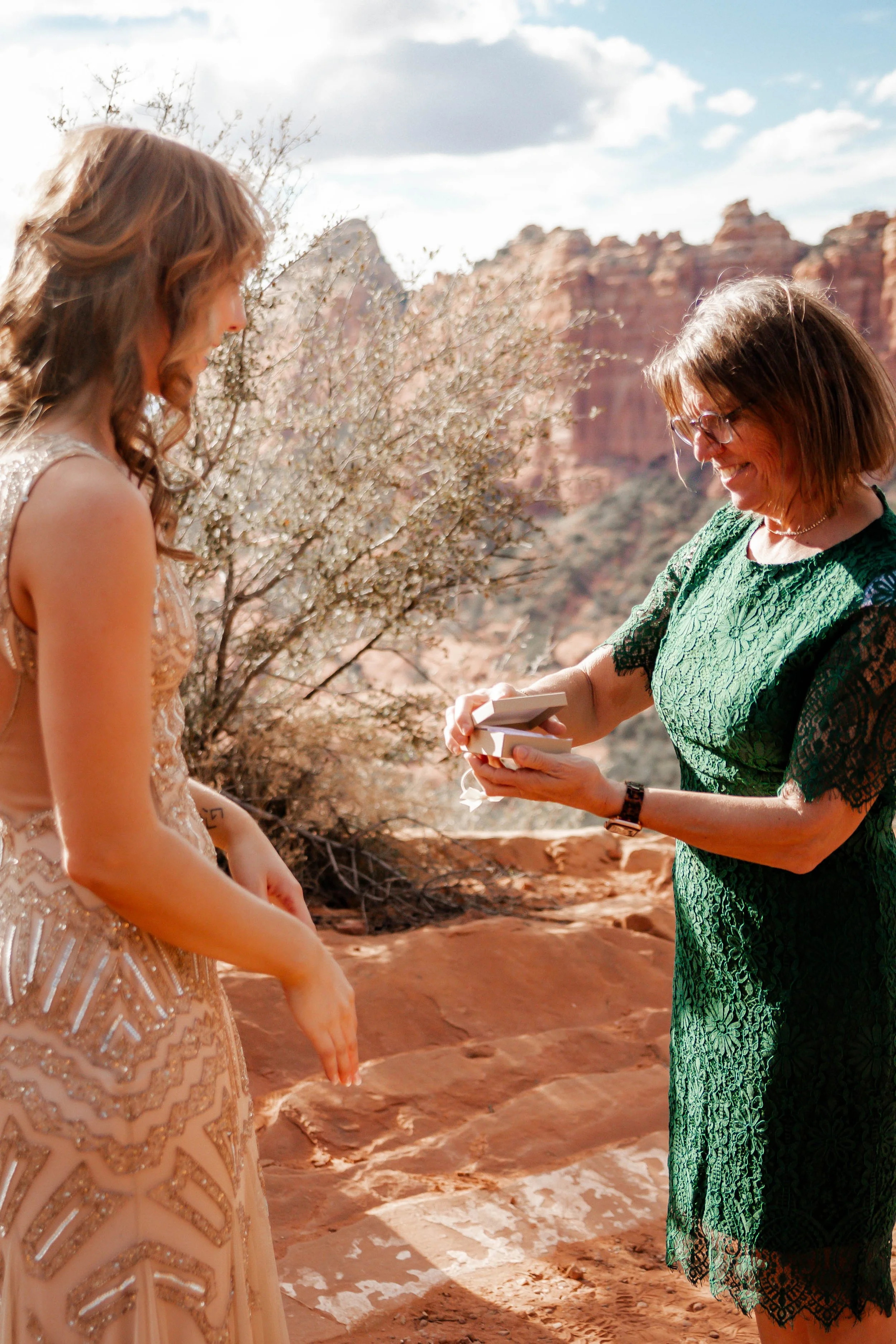 A woman in a green lace dress is presenting a ring to a younger woman wearing a light pink, beaded dress with her hands extended downward in a desert setting with red rock formations and a cloudy sky in the background.