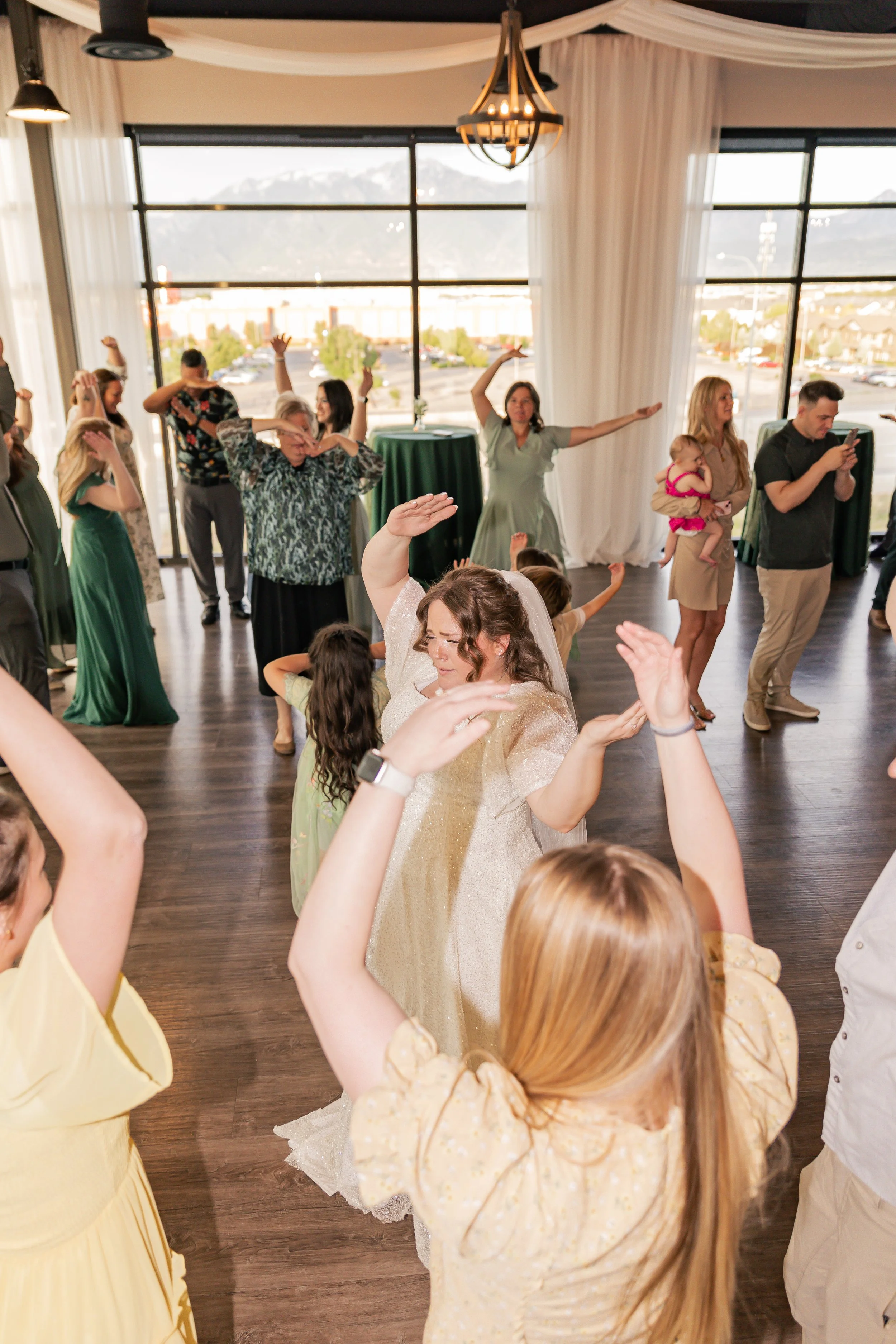 Dance floor with people dancing, a bride in a white dress at the center, and large windows showing mountains outside.