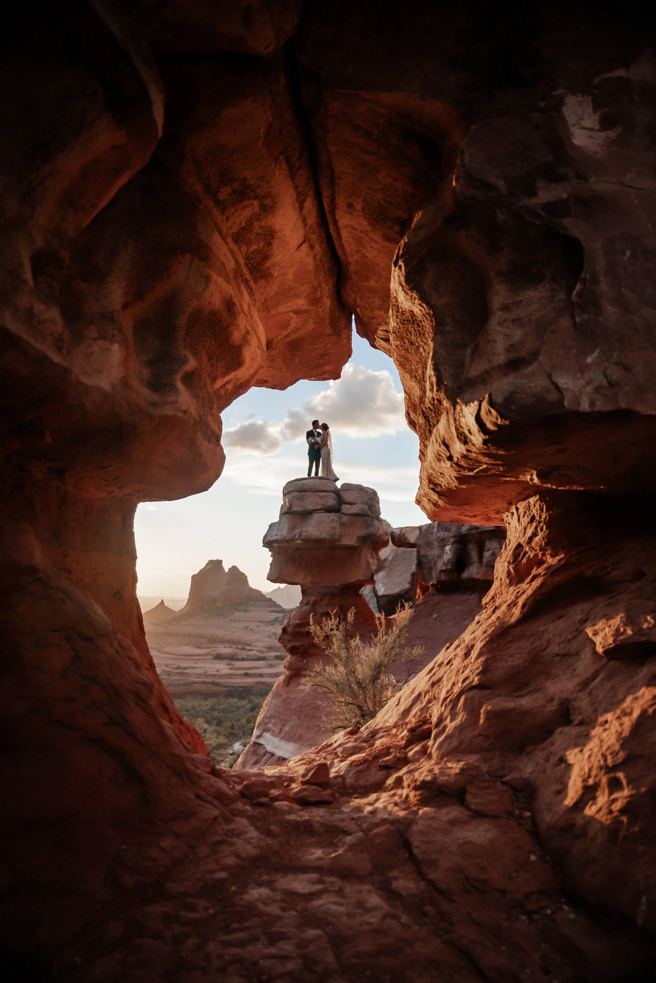 A couple standing on a rock formation, framed by a natural rock window, with a desert landscape and distant formations in the background during sunset.