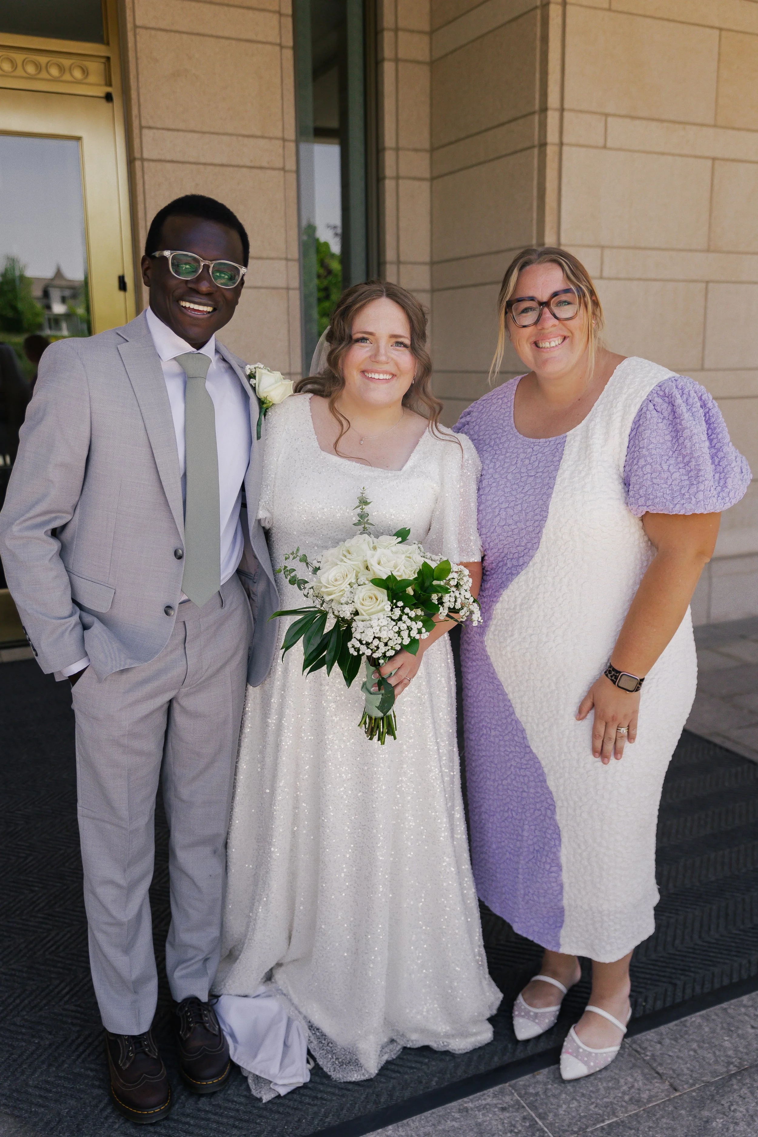 Three people standing in front of a building, during a wedding, including a groom in a light gray suit, a bride in a white dress holding a bouquet, and a woman in a white and purple dress.