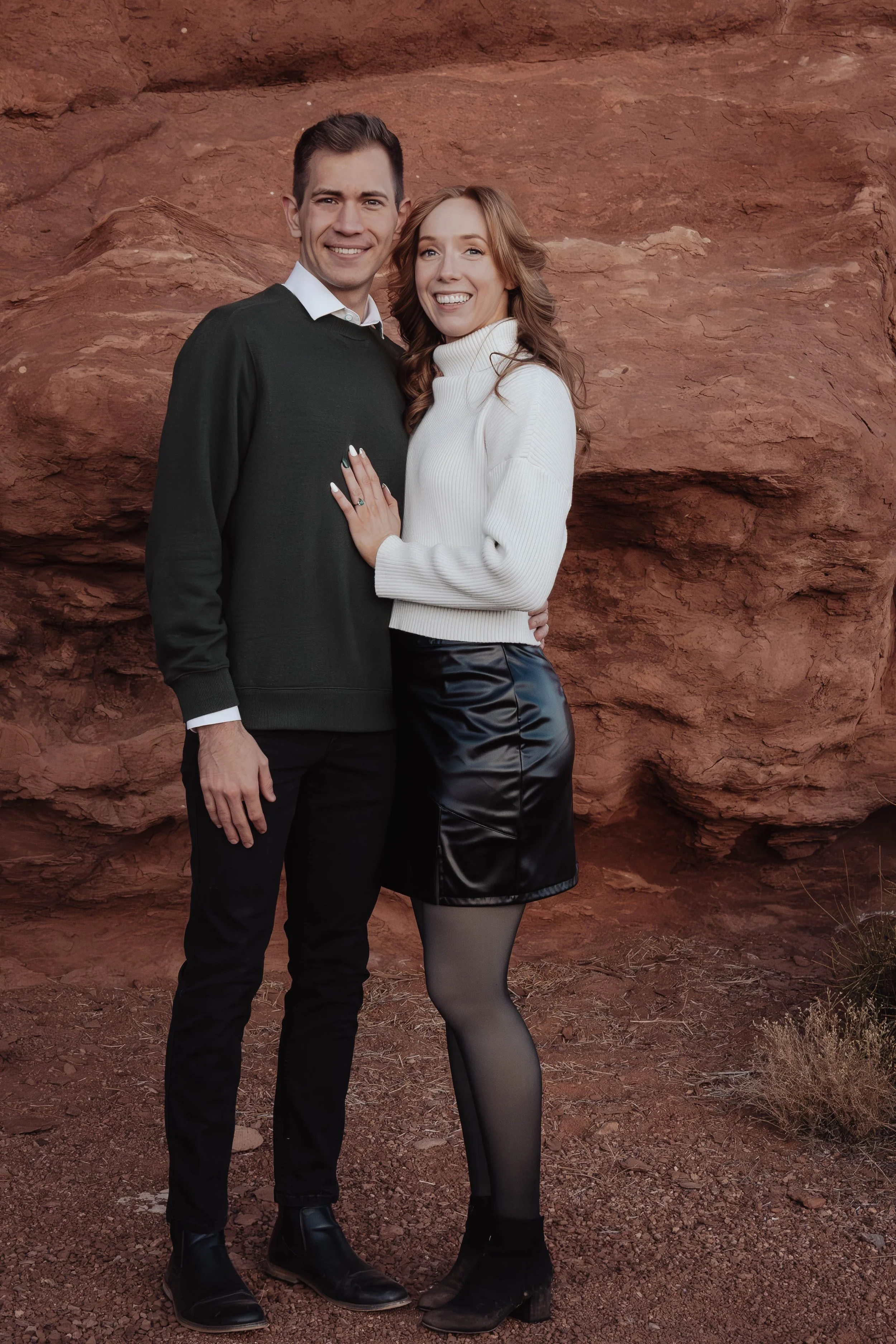 A smiling man and woman standing close together outdoors against a red rock background. The man is wearing a dark sweater, white collared shirt, black pants, and black shoes. The woman is wearing a white turtleneck sweater, black leather skirt, black