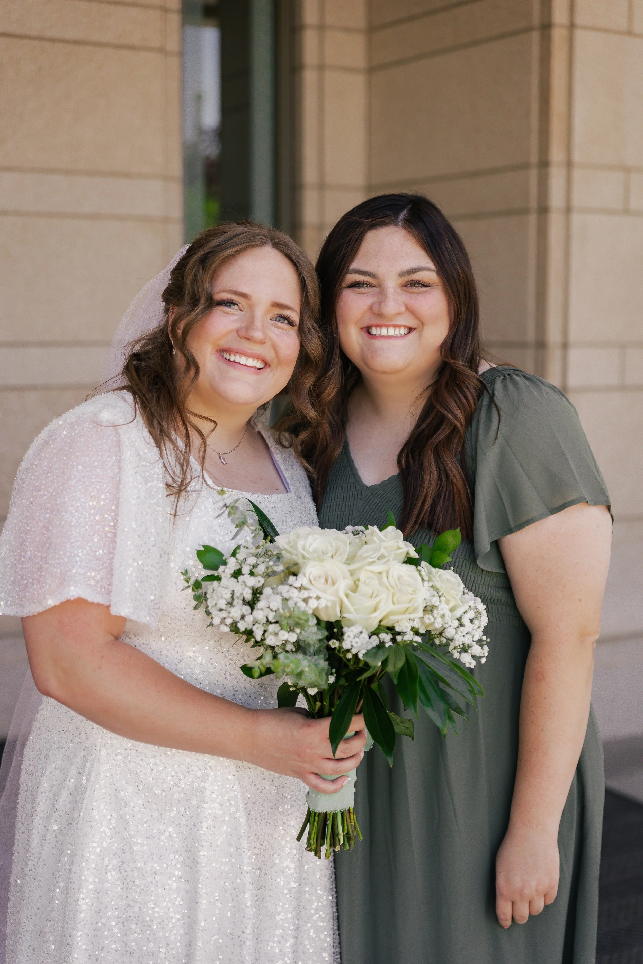 Two women smiling, with one in a wedding dress and holding a bouquet of white roses and greenery, standing outdoors in front of a beige stone wall.
