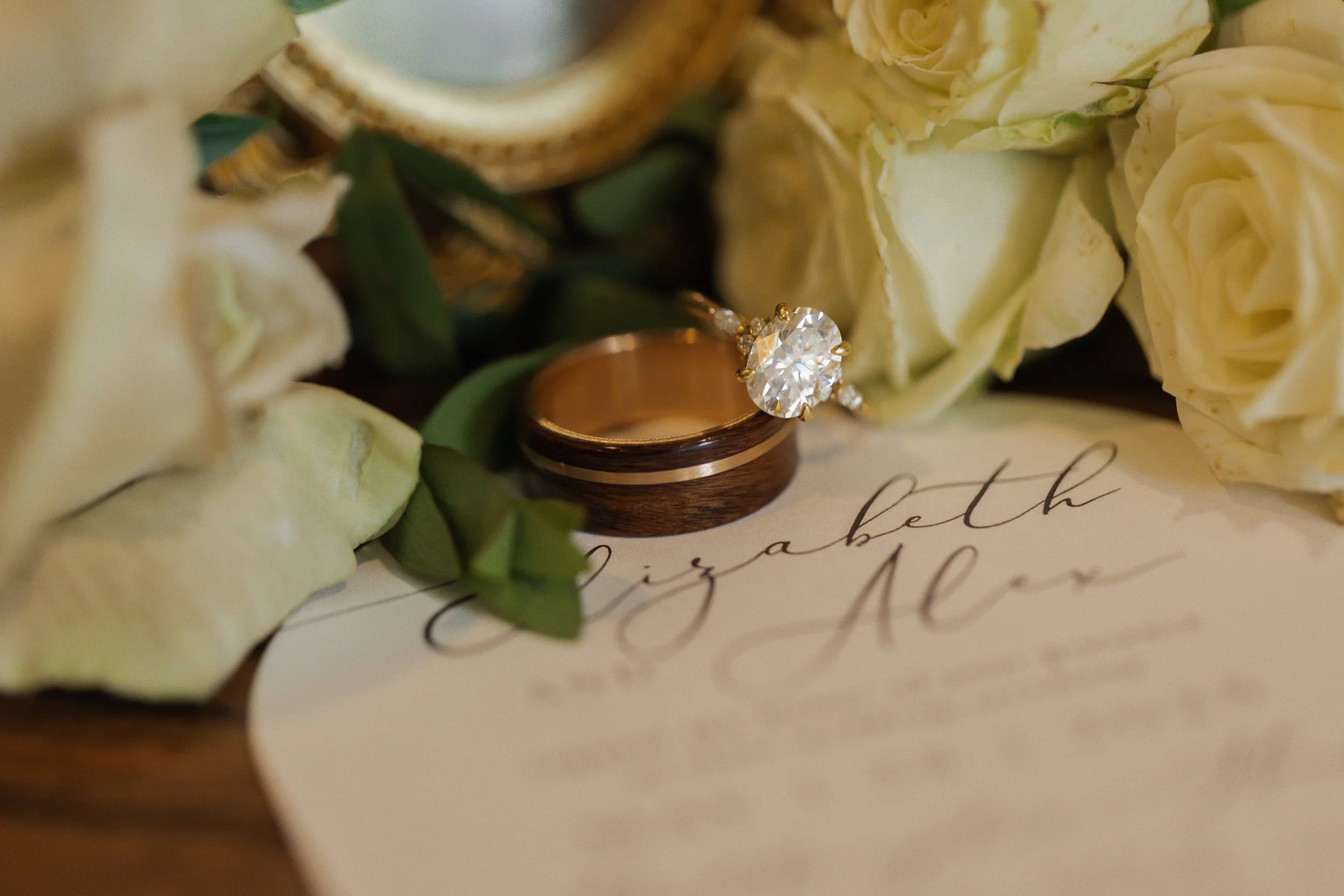 A wedding ring with a large diamond solitaire engagement ring resting on a wedding band, surrounded by white roses and greenery, and a handwritten note in elegant script.