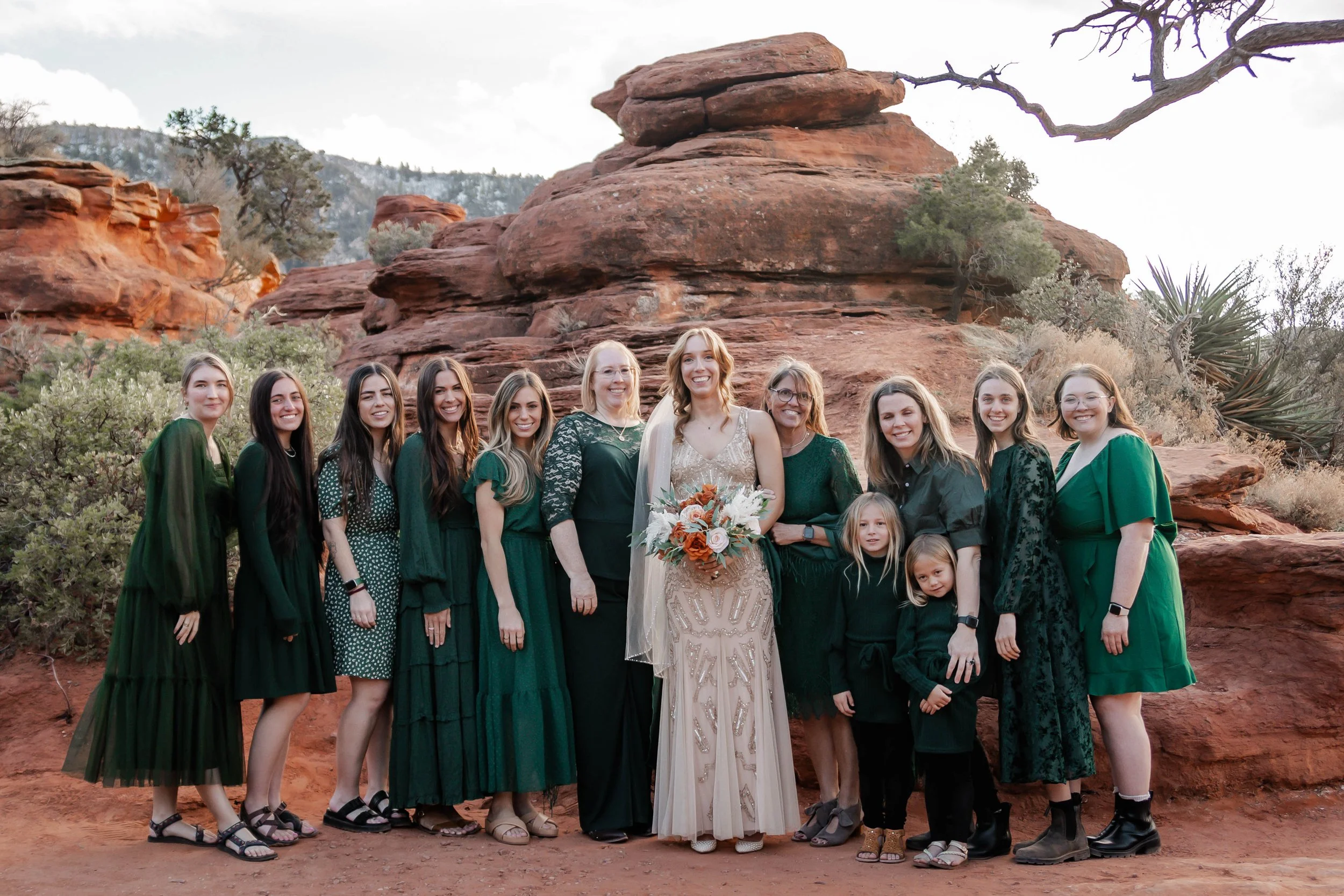A group of women and children dressed in green and black standing together on a rocky desert landscape with red rocks and sparse vegetation in the background, celebrating a wedding.