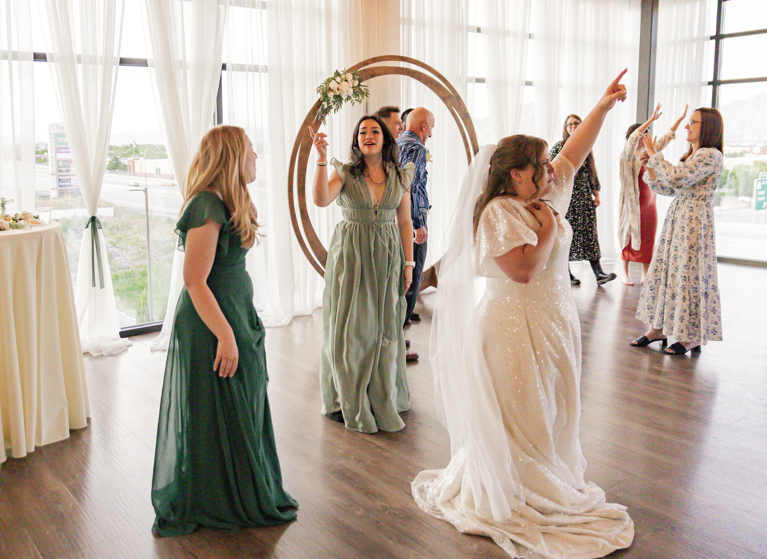 A group of women at a wedding reception dancing and celebrating, with large windows and sheer curtains in the background.
