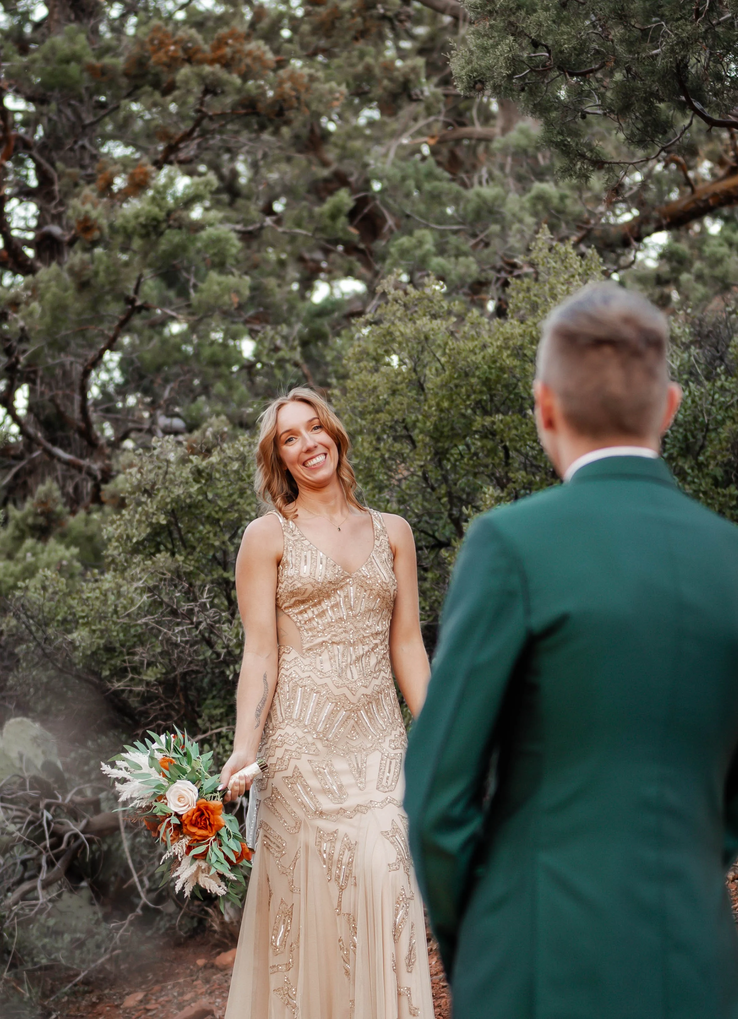 A woman in a beige, ornate dress holding a bouquet of flowers, smiling at a man in a green suit, outdoors among trees.