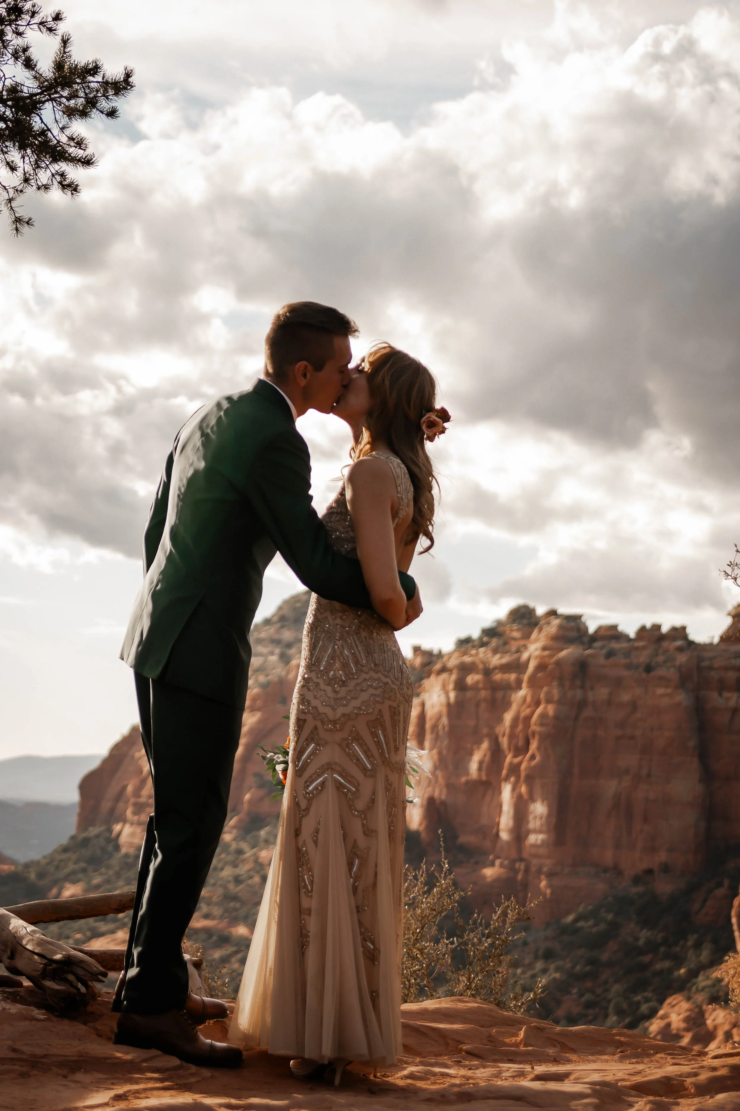 A couple in wedding attire sharing a kiss against a desert mountainscape, with dramatic clouds overhead.