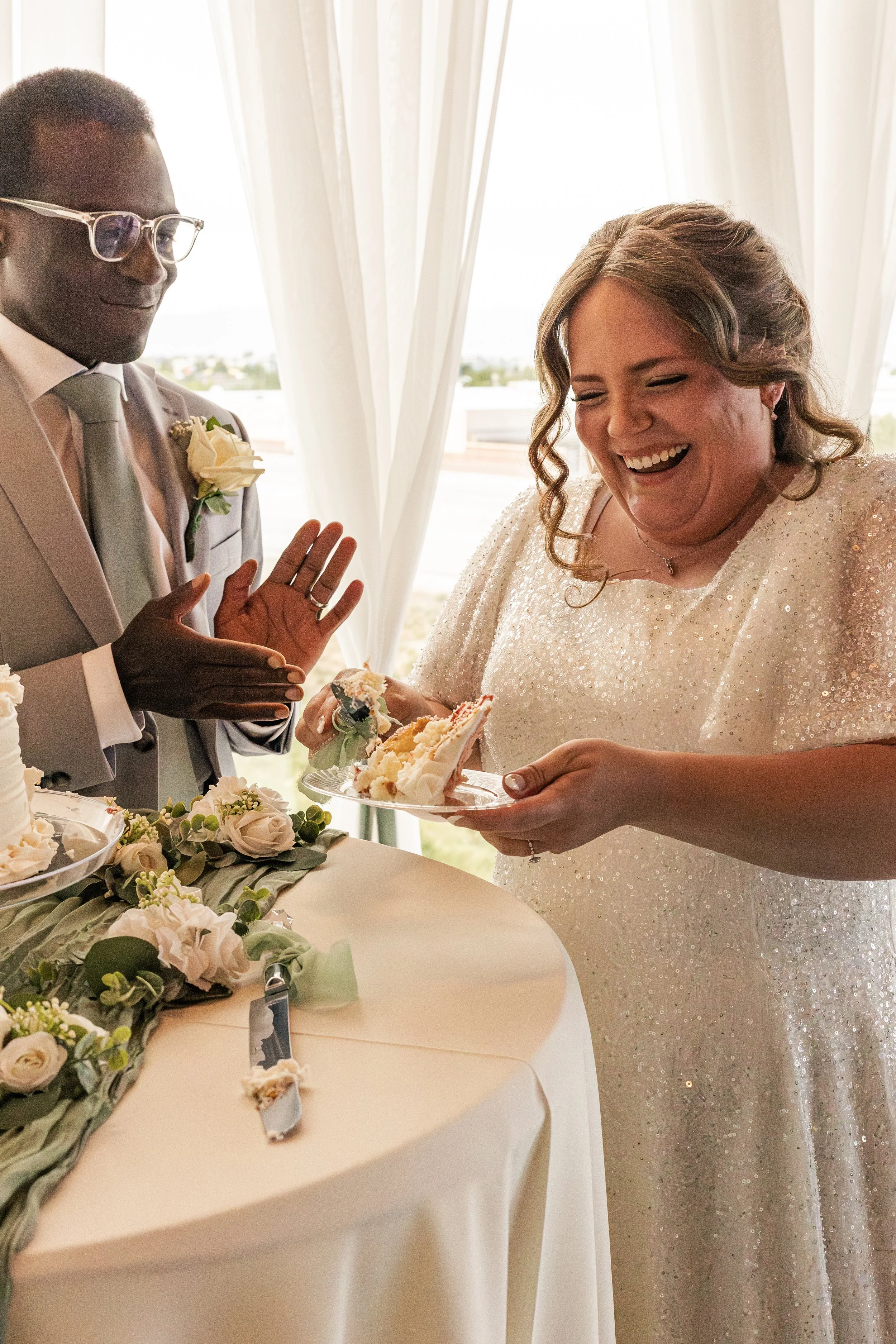 A bride and groom at a wedding reception, the bride is laughing and holding a plate with a slice of cake, while the groom is clapping his hands.