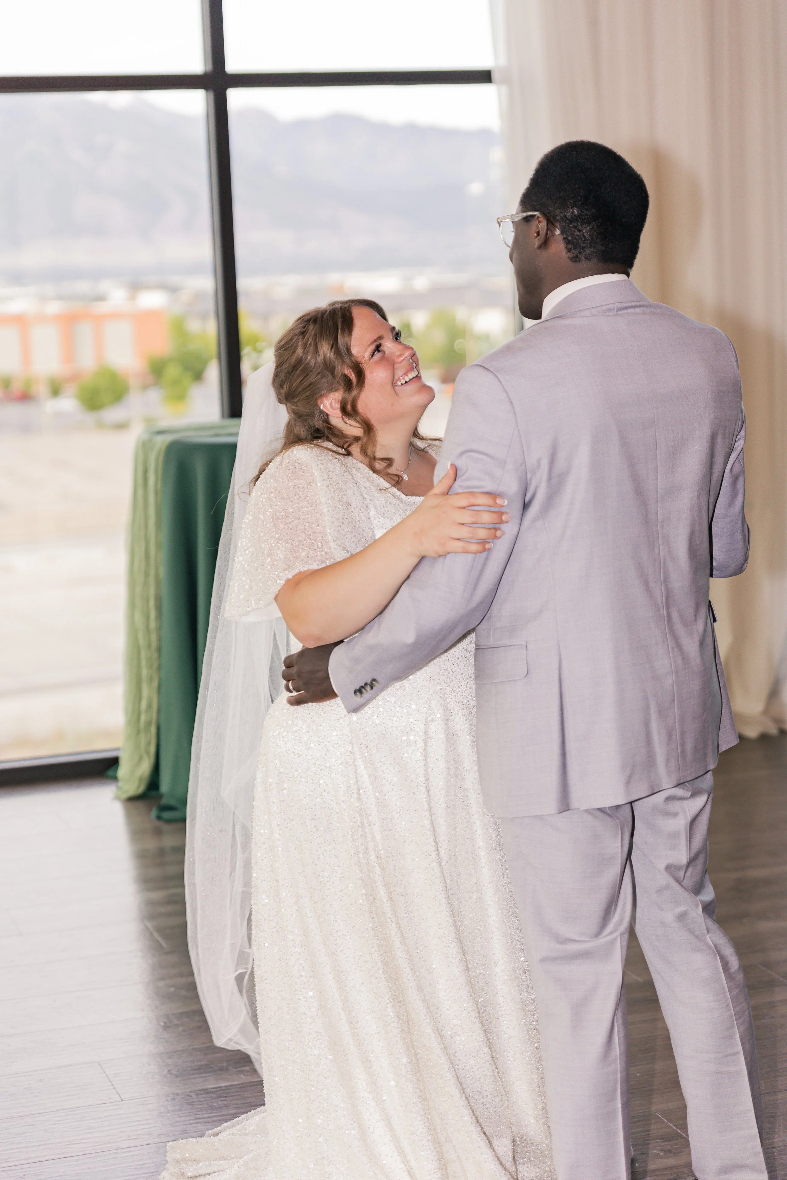 A bride and groom dancing indoors during their wedding reception, with large windows and mountain scenery in the background.