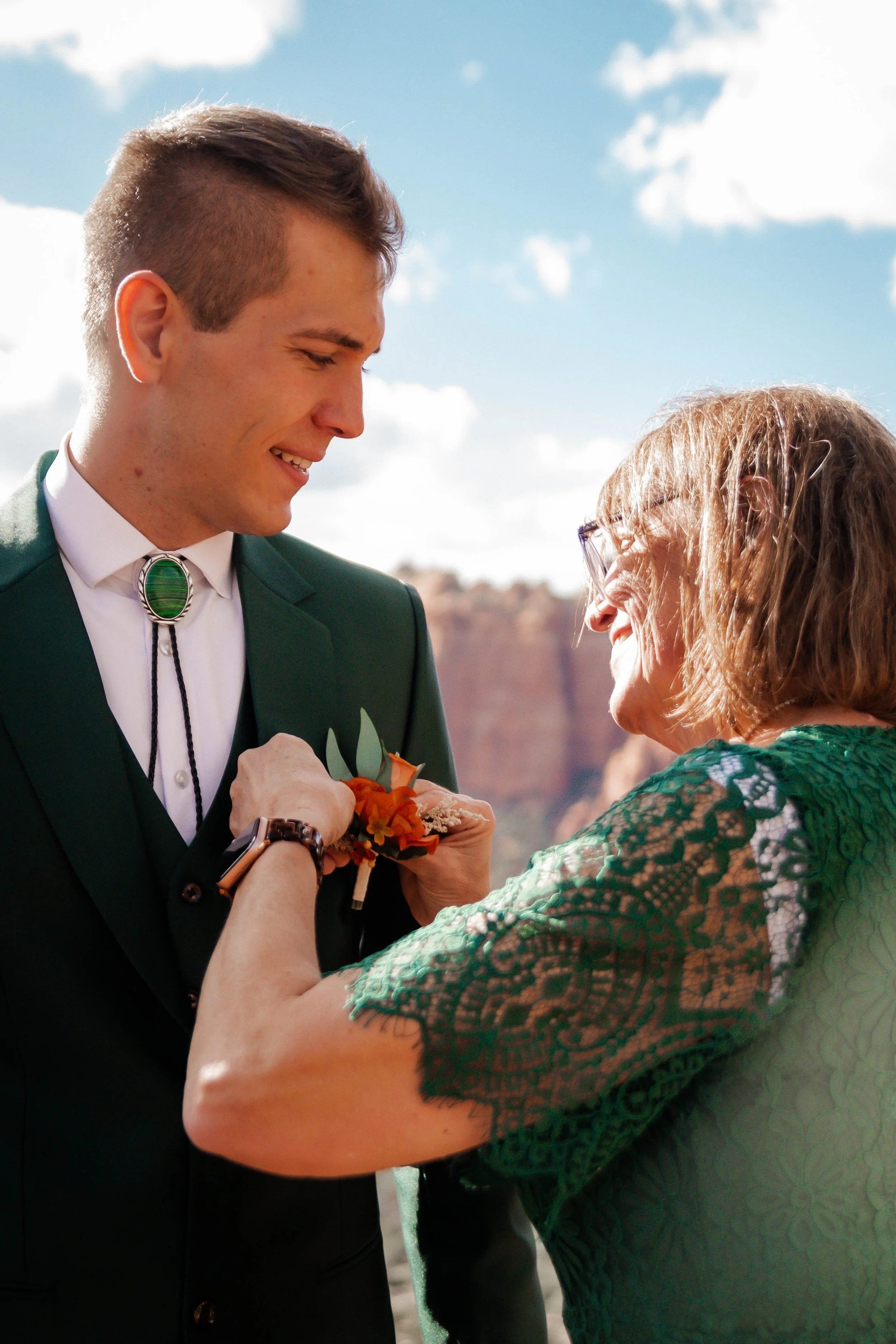 A young man wearing a dark green suit and bolo tie smiling as an older woman with glasses pins a carnation corsage to his lapel outdoors with red rock formations and a partly cloudy sky in the background.