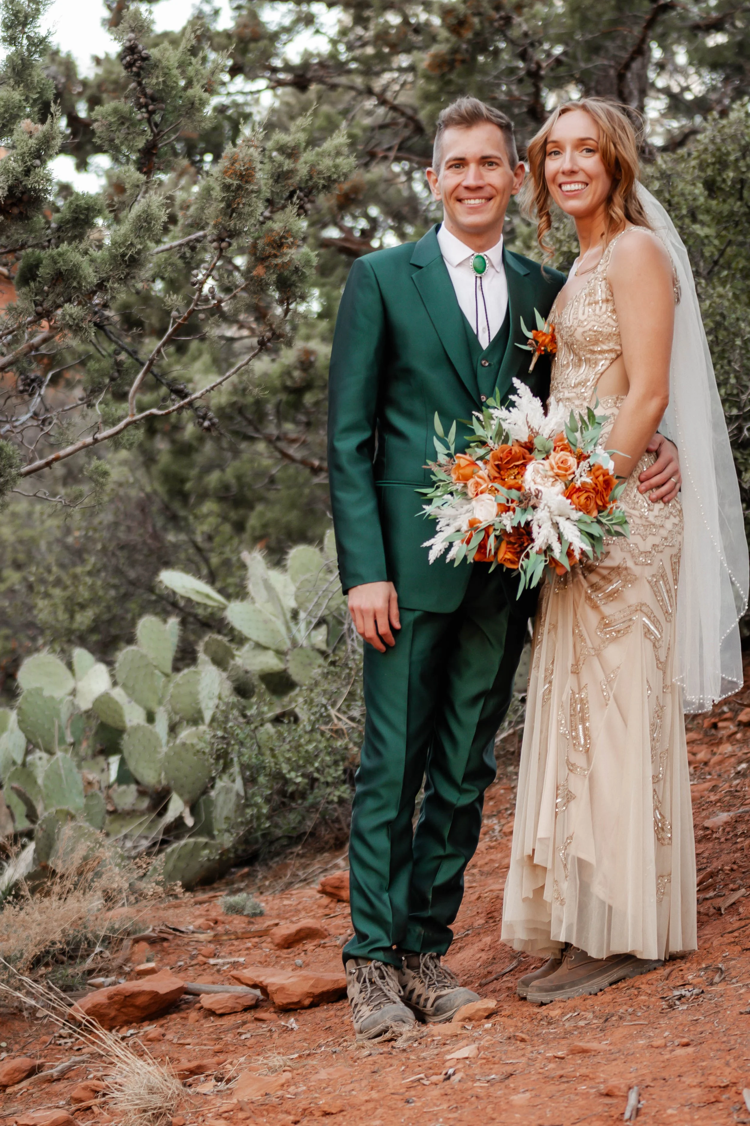 A bride and groom standing outdoors surrounded by trees and desert plants, smiling at the camera. The groom is wearing a green suit with a bolo tie, and the bride is dressed in a beige, beaded gown holding a bouquet of orange, white, and green flower