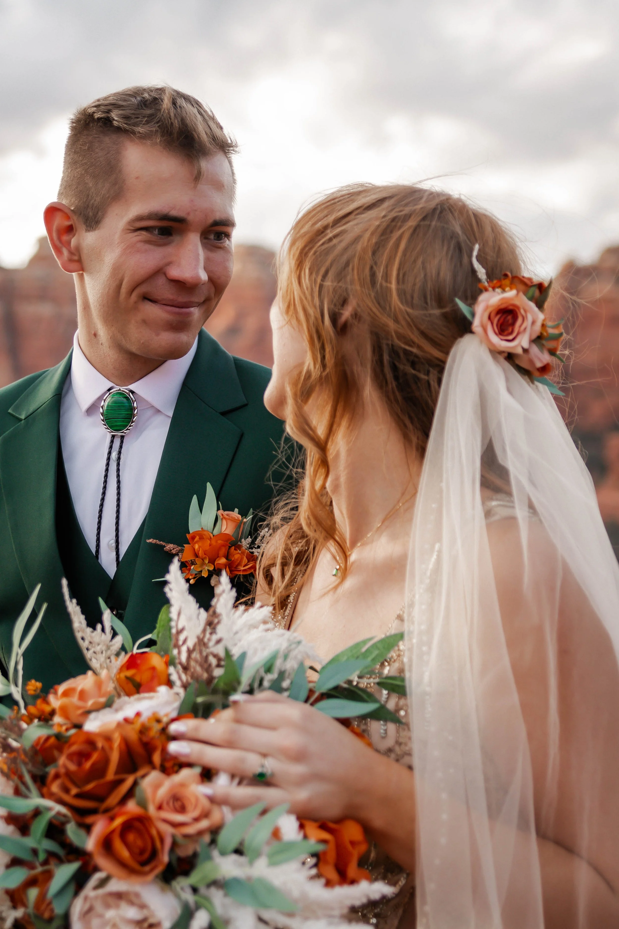 A groom and bride on their wedding day outdoors, with background of red rocks. The groom is in a green suit with a bolo tie, and the bride has red hair, a floral hairpiece, and a veil. She is holding a bouquet of orange and pink roses with greenery.