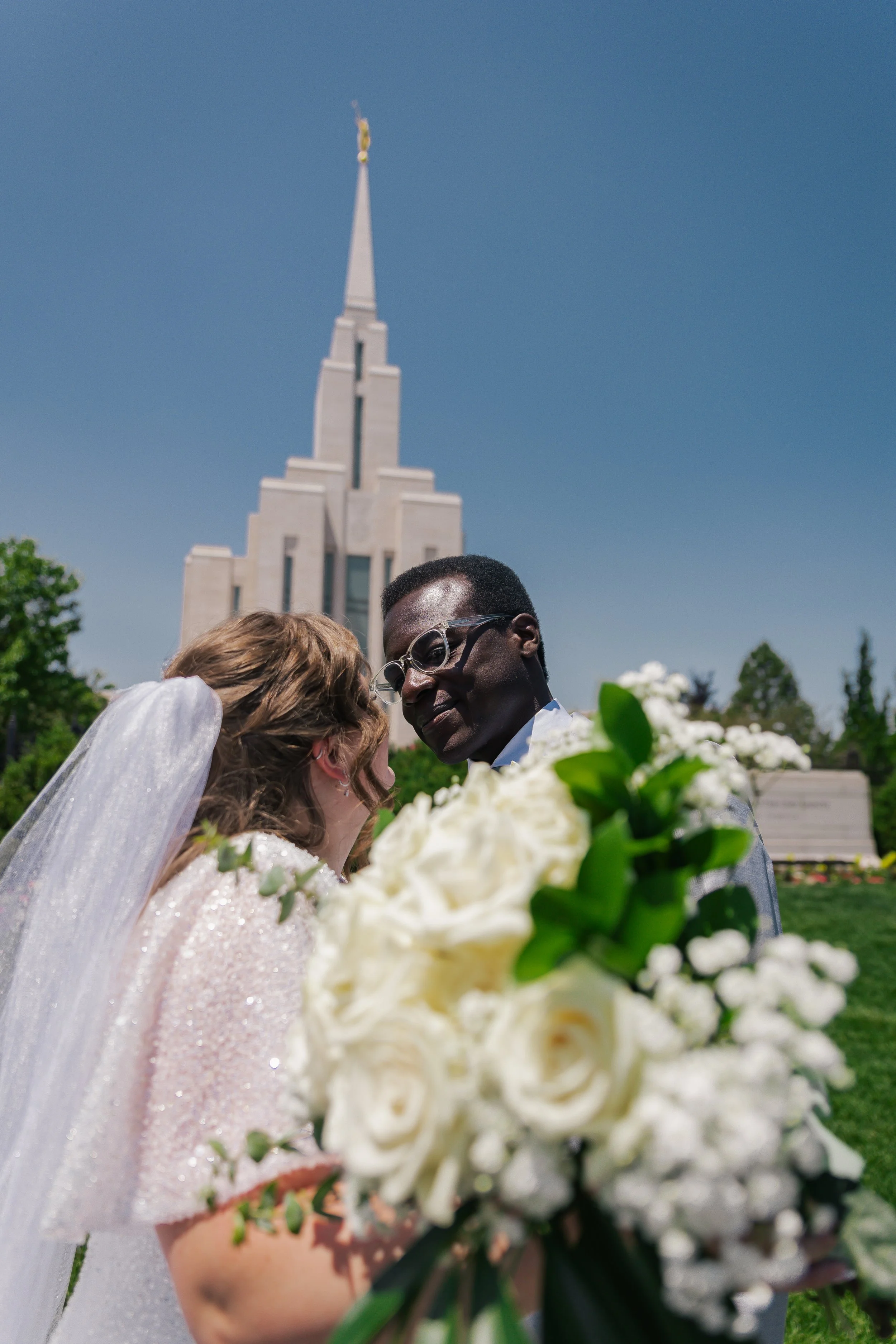A bride and groom stand close together, smiling, in front of oquirrh mountain temple during their wedding ceremony. The bride has a veil and is holding a bouquet of white roses, and the groom is wearing glasses and a suit.